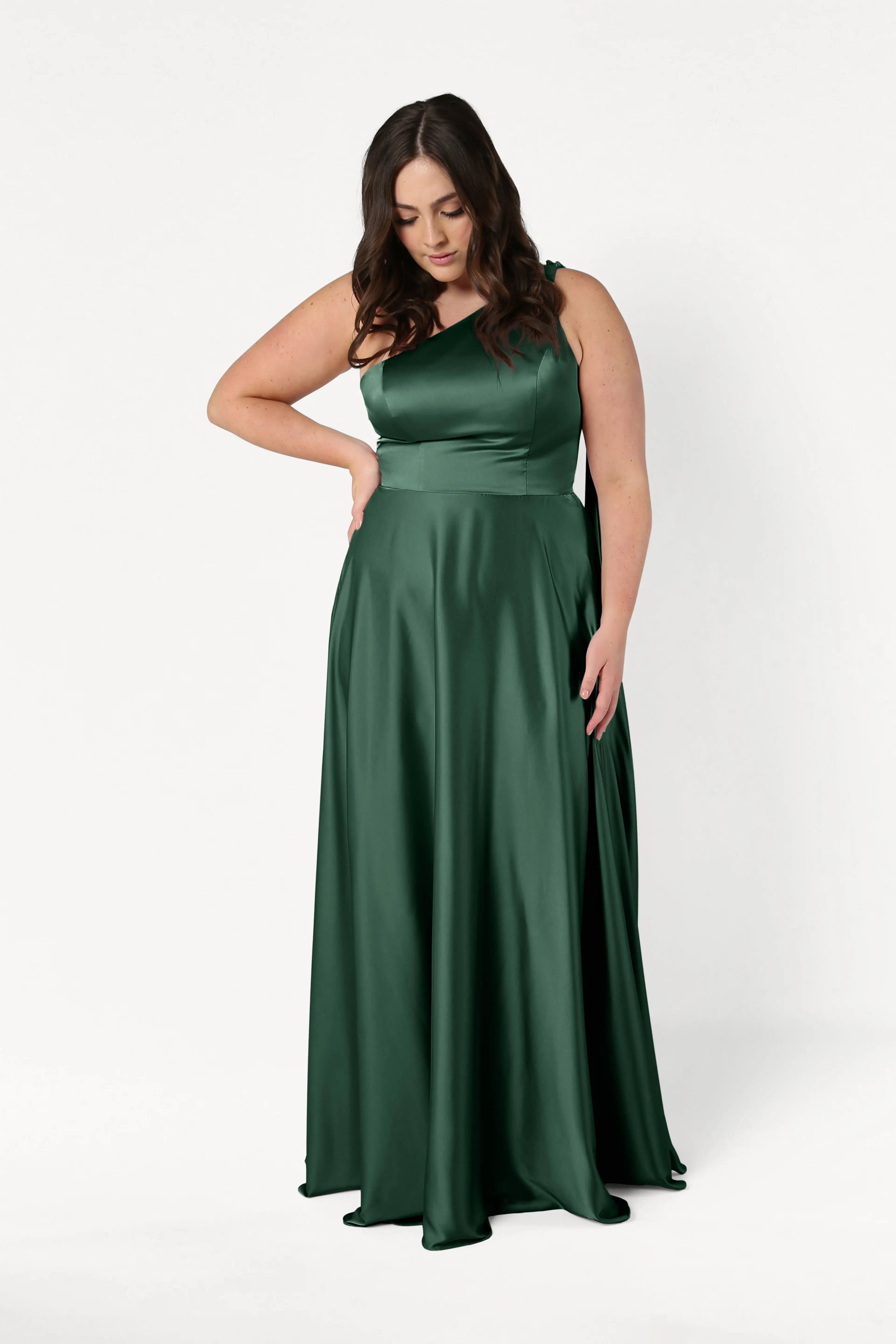 A woman in the Lyra Bridesmaid Dress in Olive Grove Satin by TH&TH stands against a plain white background, looking down with one hand on her hip. This elegant satin gown features a size-inclusive, one-shoulder design.