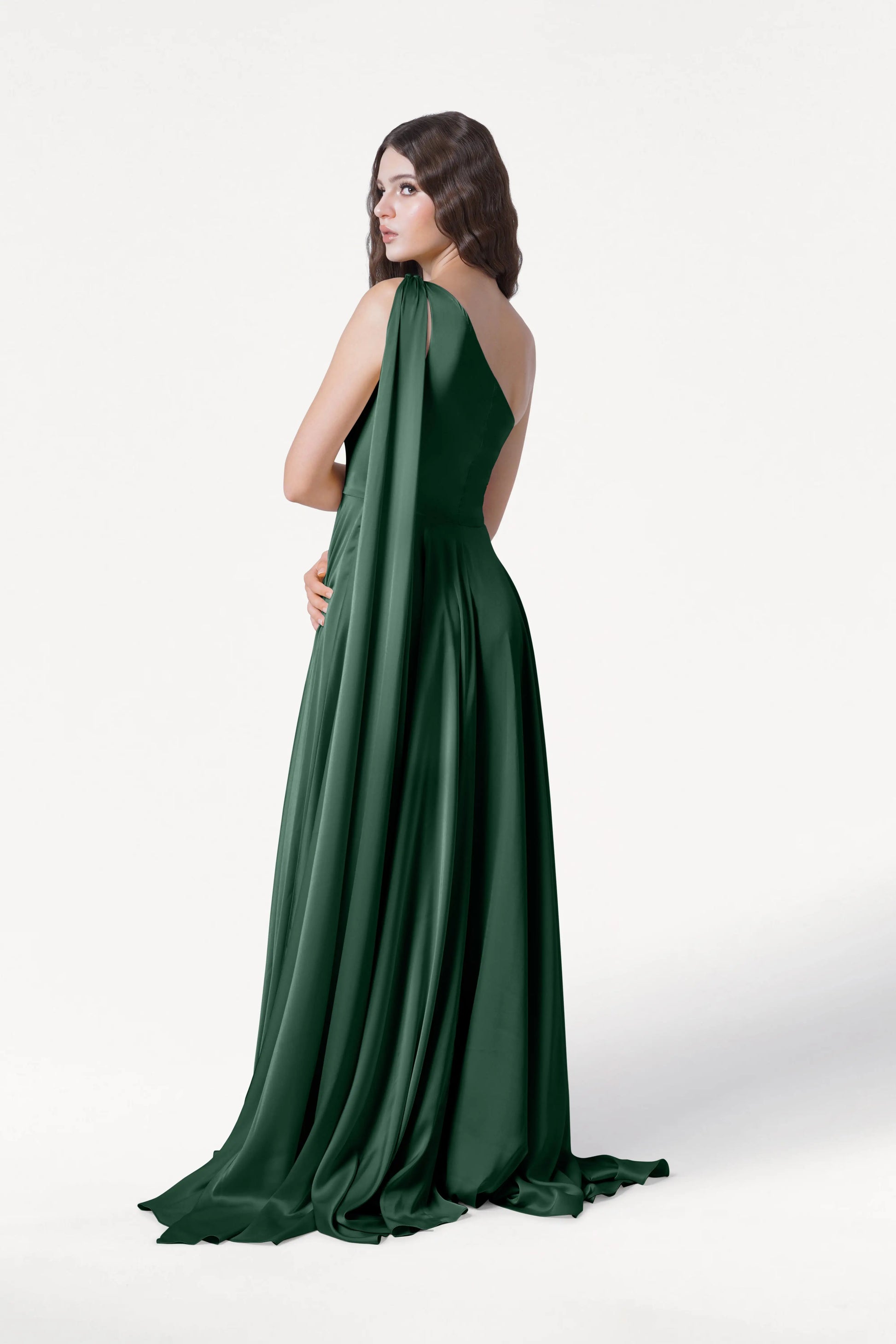 A woman with long brown hair wears the TH&TH Lyra Bridesmaid Dress in Olive Grove Satin, a floor-length, one-shoulder gown with draped fabric. She looks over her shoulder against a plain white background.