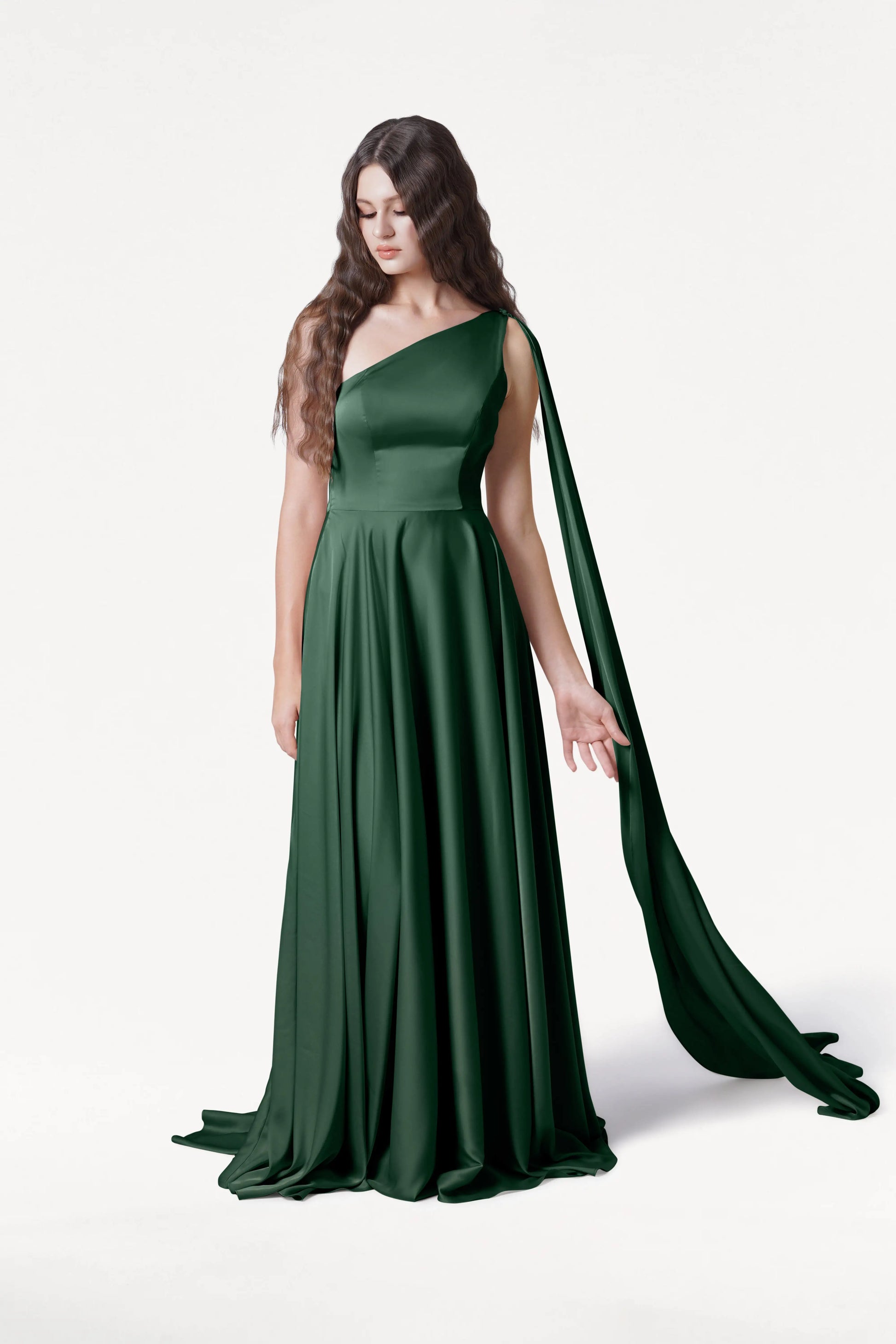 A woman with long wavy brown hair wears the TH&TH Lyra Bridesmaid Dress in Olive Grove Satin, a floor-length, size-inclusive one-shoulder gown with a flowing arm drape. She stands against a plain white background, gazing downward.
