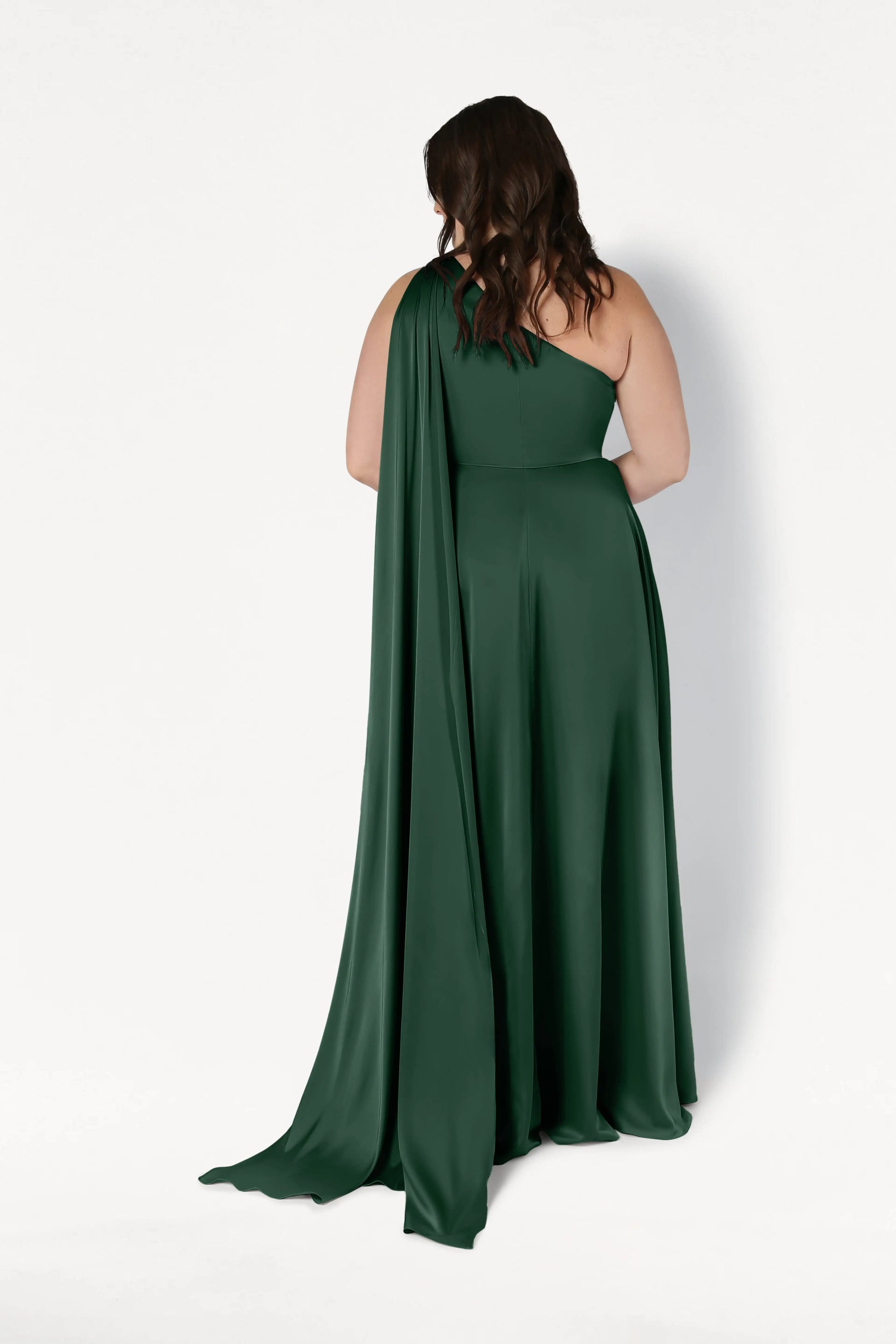 A woman with wavy brown hair faces away, wearing the TH&TH Lyra Bridesmaid Dress in Olive Grove Satin, a dark green, size-inclusive one-shoulder gown with a long drape. The background is plain white.