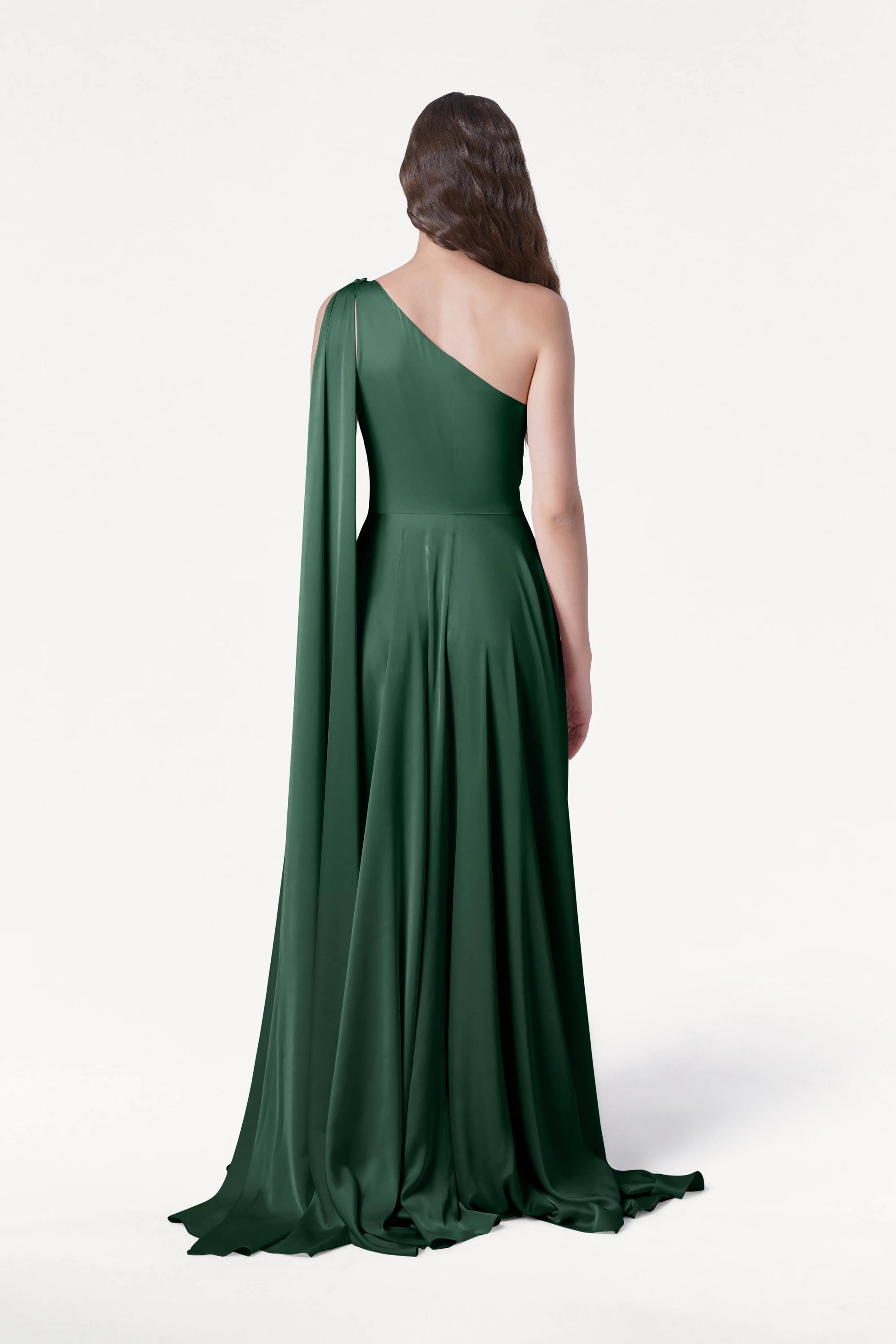 A woman with long brown hair stands facing away, wearing the size-inclusive TH&TH Lyra Bridesmaid Dress in Olive Grove Satin, a floor-length one-shoulder gown with a flowing drape, set against a plain white background.