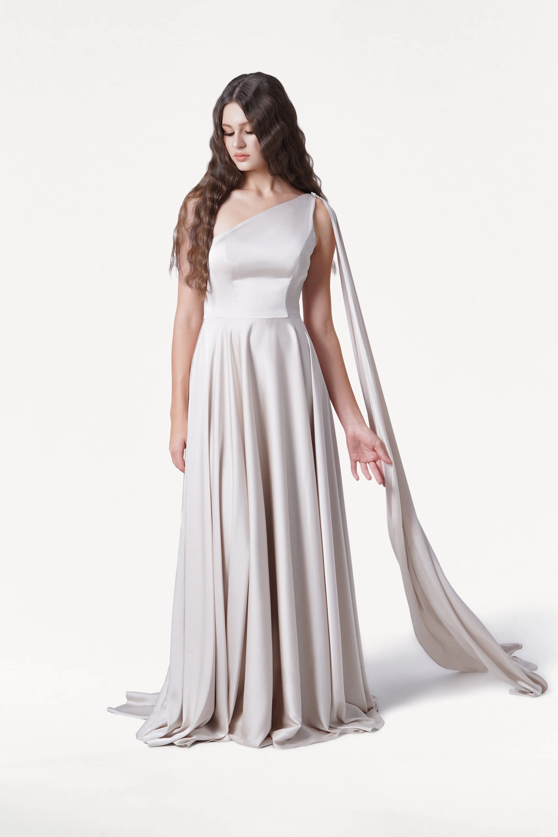 A woman with long, wavy brown hair wears the size-inclusive Lyra Bridesmaid Dress in Oyster Satin by TH&TH, featuring a flowing one-shoulder design with draped fabric. She stands against a white background, looking down with one hand slightly raised.