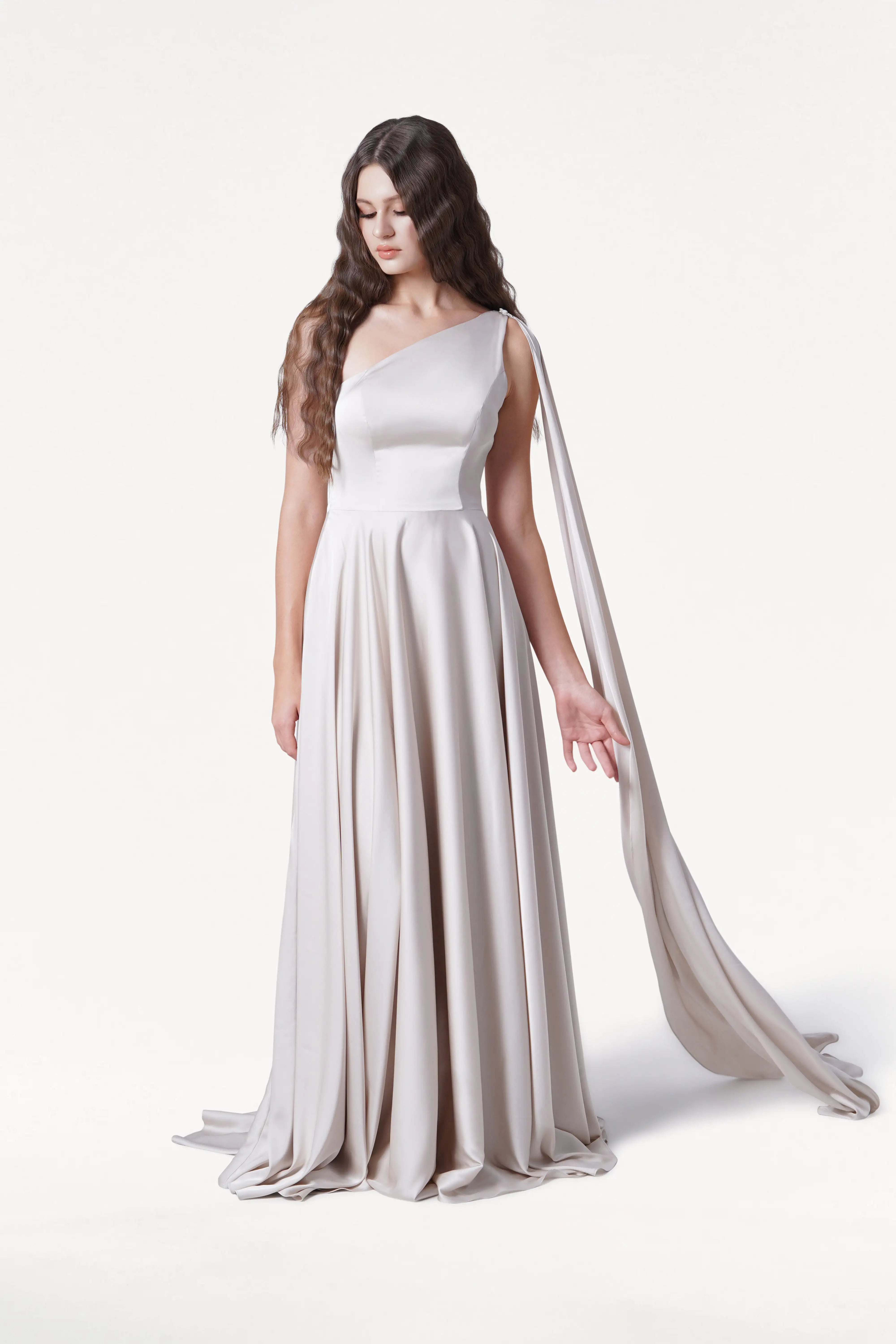 A woman with long, wavy brown hair wears the size-inclusive Lyra Bridesmaid Dress in Oyster Satin by TH&TH, featuring a flowing one-shoulder design with draped fabric. She stands against a white background, looking down with one hand slightly raised.