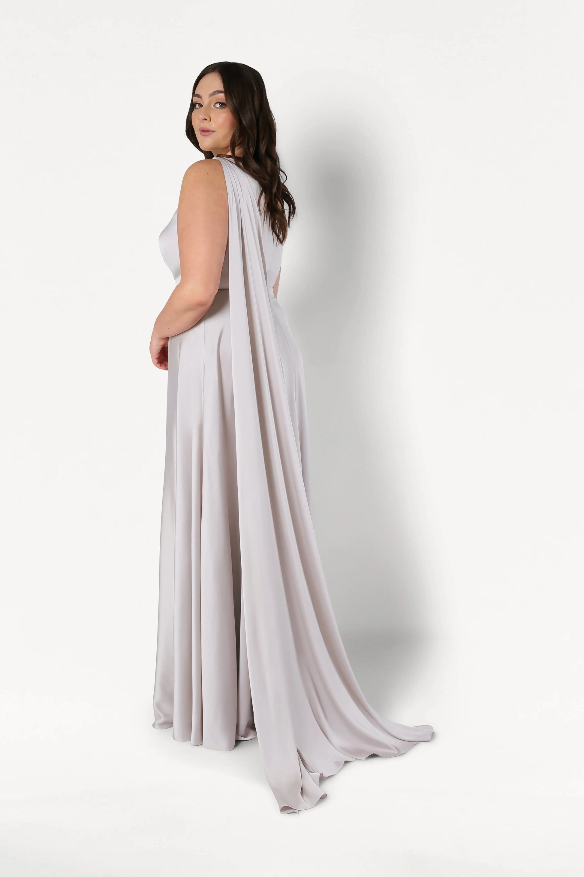 A woman with long brown hair models the TH&TH Lyra Bridesmaid Dress in Oyster Satin, a size-inclusive, floor-length gown with a dramatic draped back, standing sideways and looking over her shoulder against a white background.