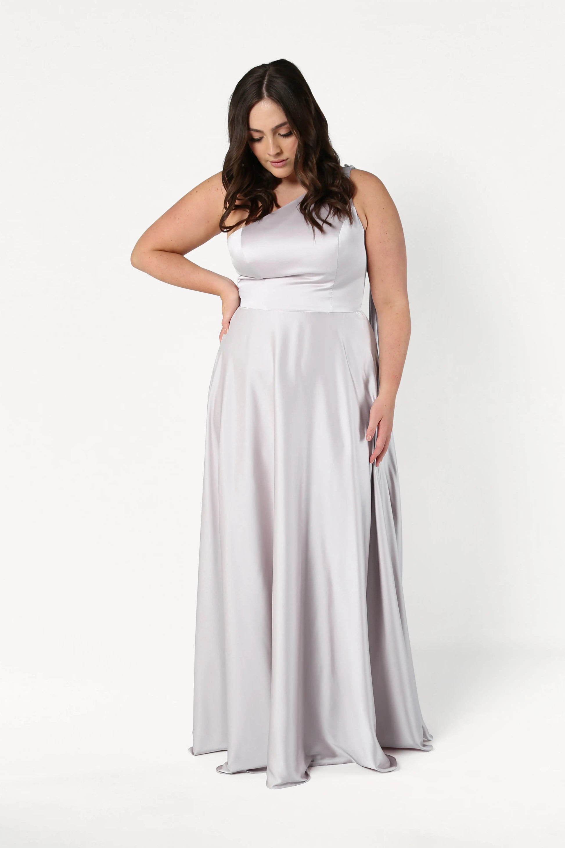 A woman with long dark hair models the size-inclusive Lyra Bridesmaid Dress in Oyster Satin by TH&TH, standing against a plain white background, looking down with one hand on her hip and the other by her side.