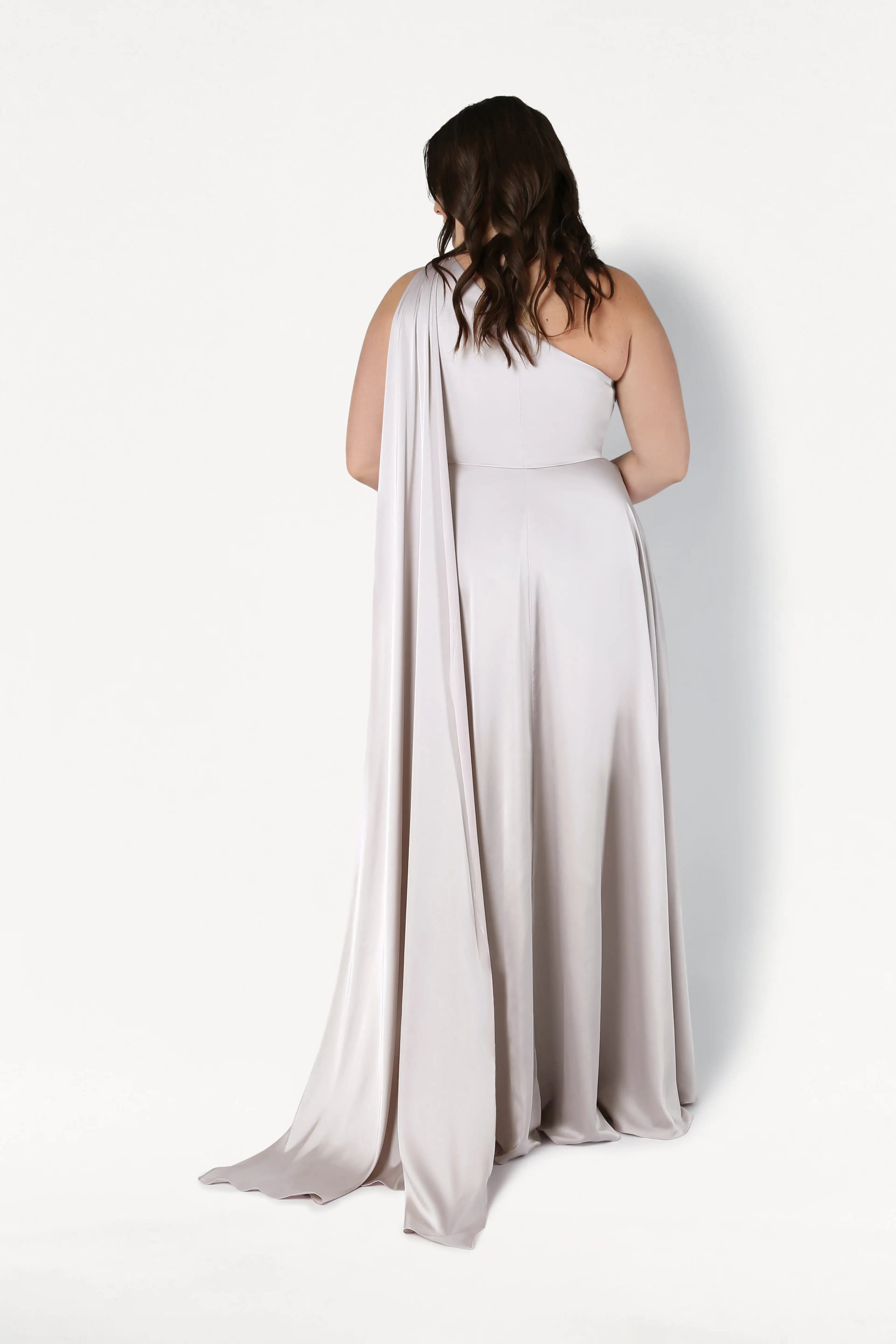 A woman with wavy brown hair models the size-inclusive Lyra Bridesmaid Dress in Oyster Satin by TH&TH—a light gray, one-shoulder gown with a long draped panel—posing against a white background to showcase its elegant satin design.