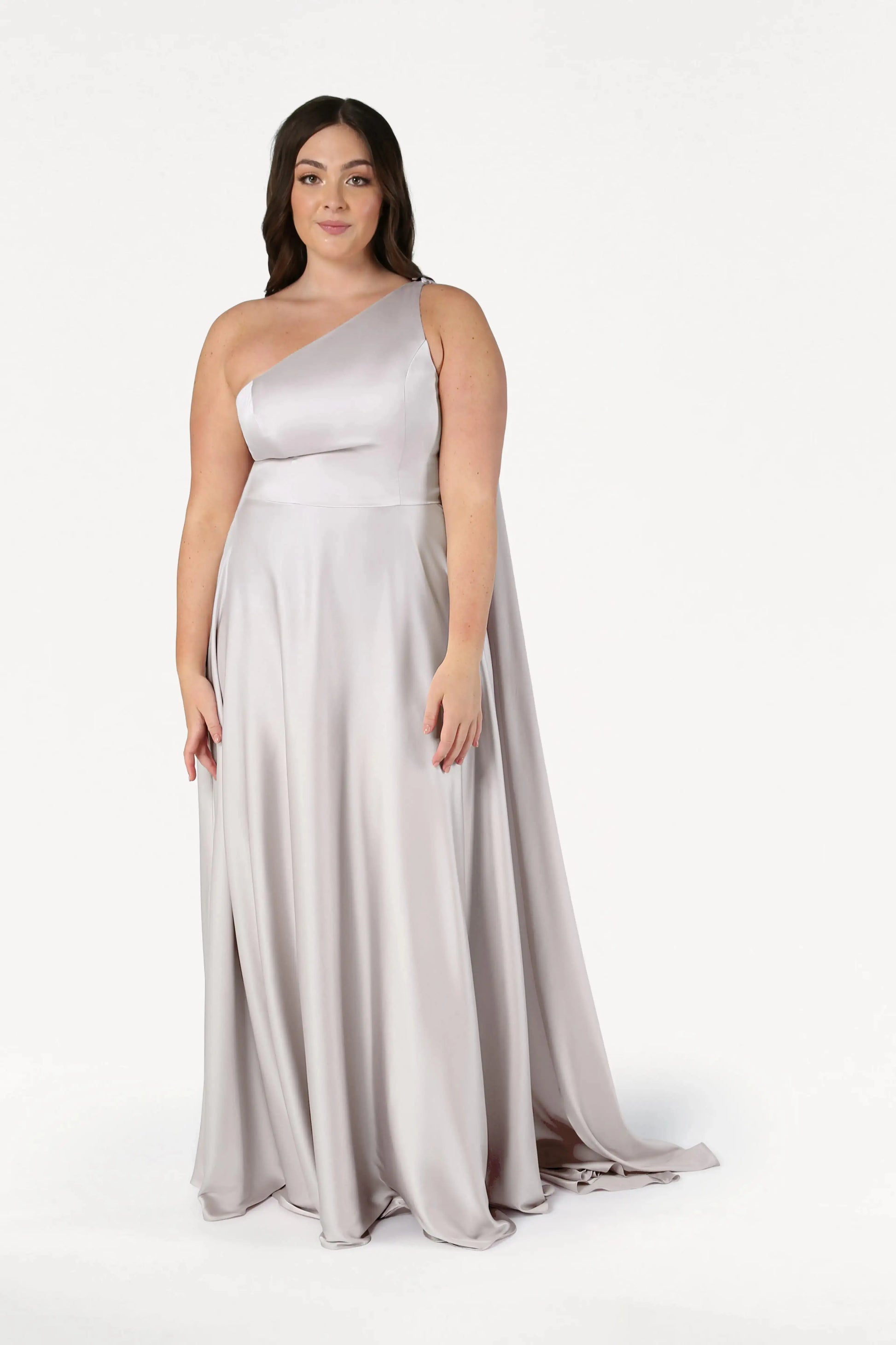 A woman with long, dark hair and a confident expression models the TH&TH Lyra Bridesmaid Dress in size-inclusive Oyster Satin with one shoulder strap, standing against a plain white background.