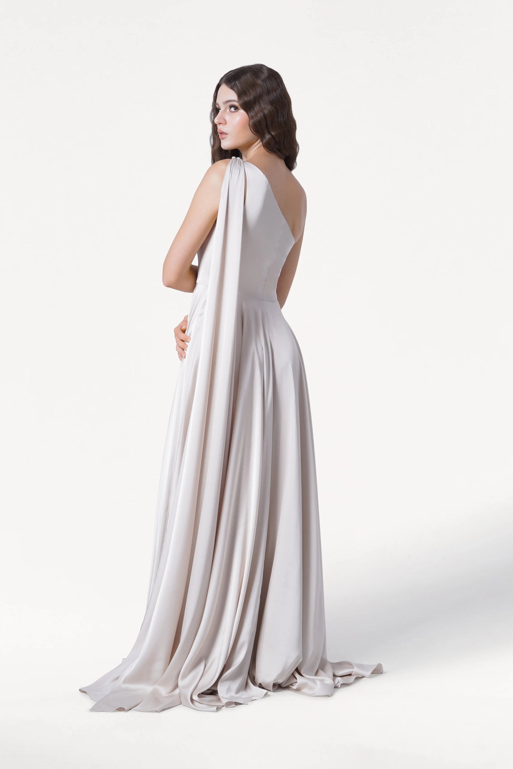 A woman with long dark hair models the Lyra Bridesmaid Dress in Oyster Satin by TH&TH, a size-inclusive one-shoulder gown that drapes to the floor. She poses against a plain white background, looking over her shoulder.
