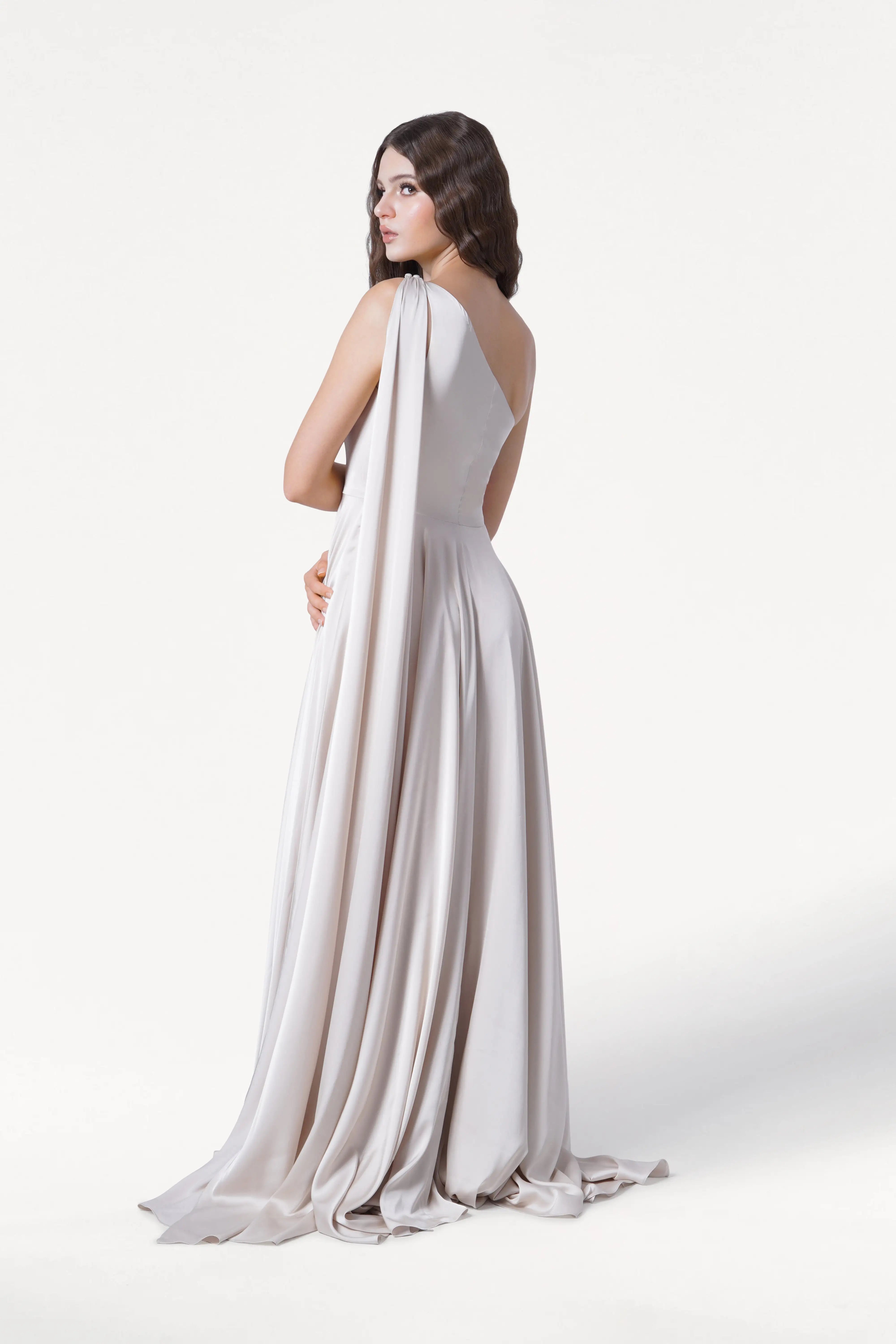 A woman with long dark hair models the Lyra Bridesmaid Dress in Oyster Satin by TH&TH, a size-inclusive one-shoulder gown that drapes to the floor. She poses against a plain white background, looking over her shoulder.