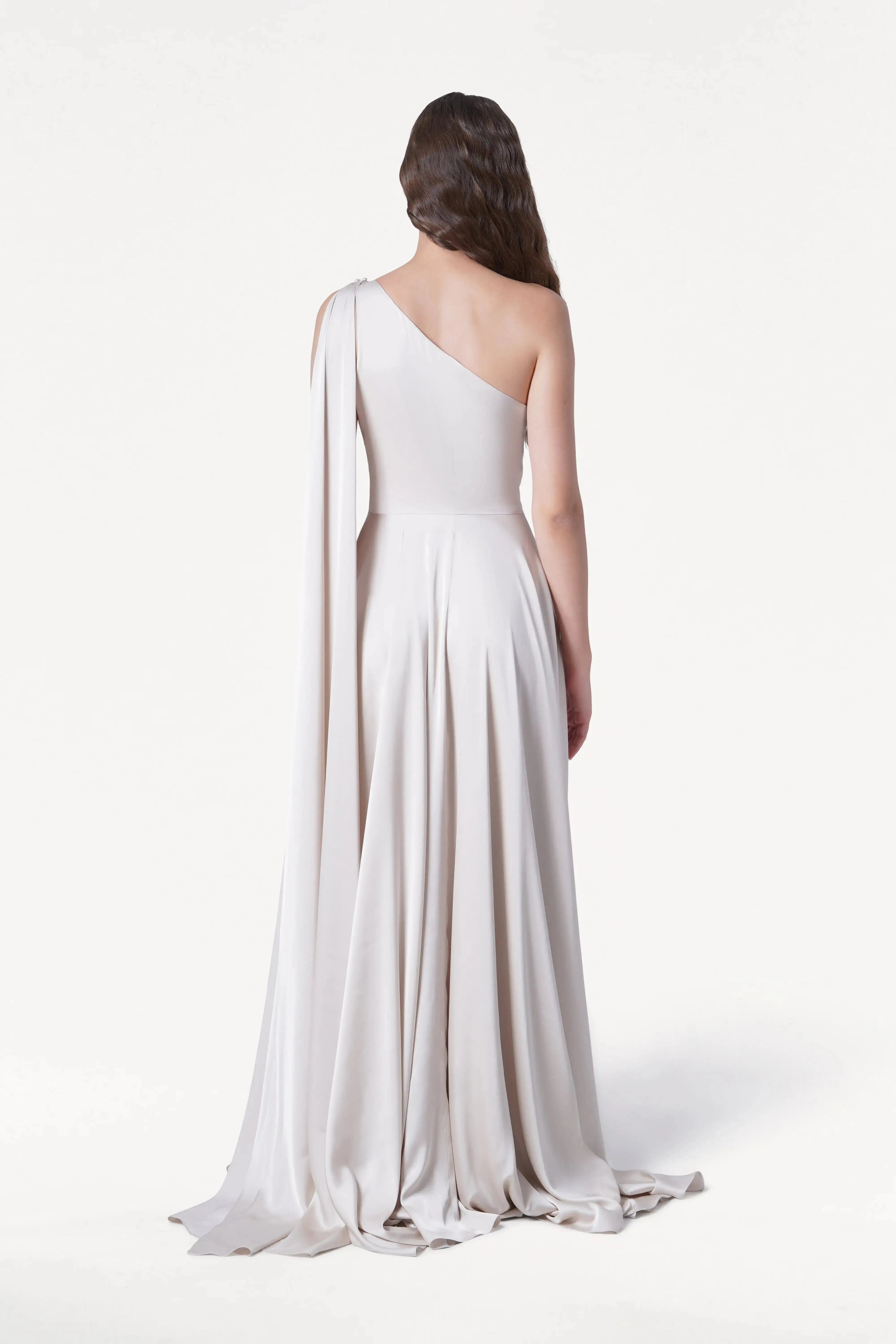 A woman with wavy brown hair stands facing away in the Lyra Bridesmaid Dress in Oyster Satin by TH&TH—a size-inclusive, one-shoulder gown with a draped sleeve and floor-length skirt—set against a plain white background.