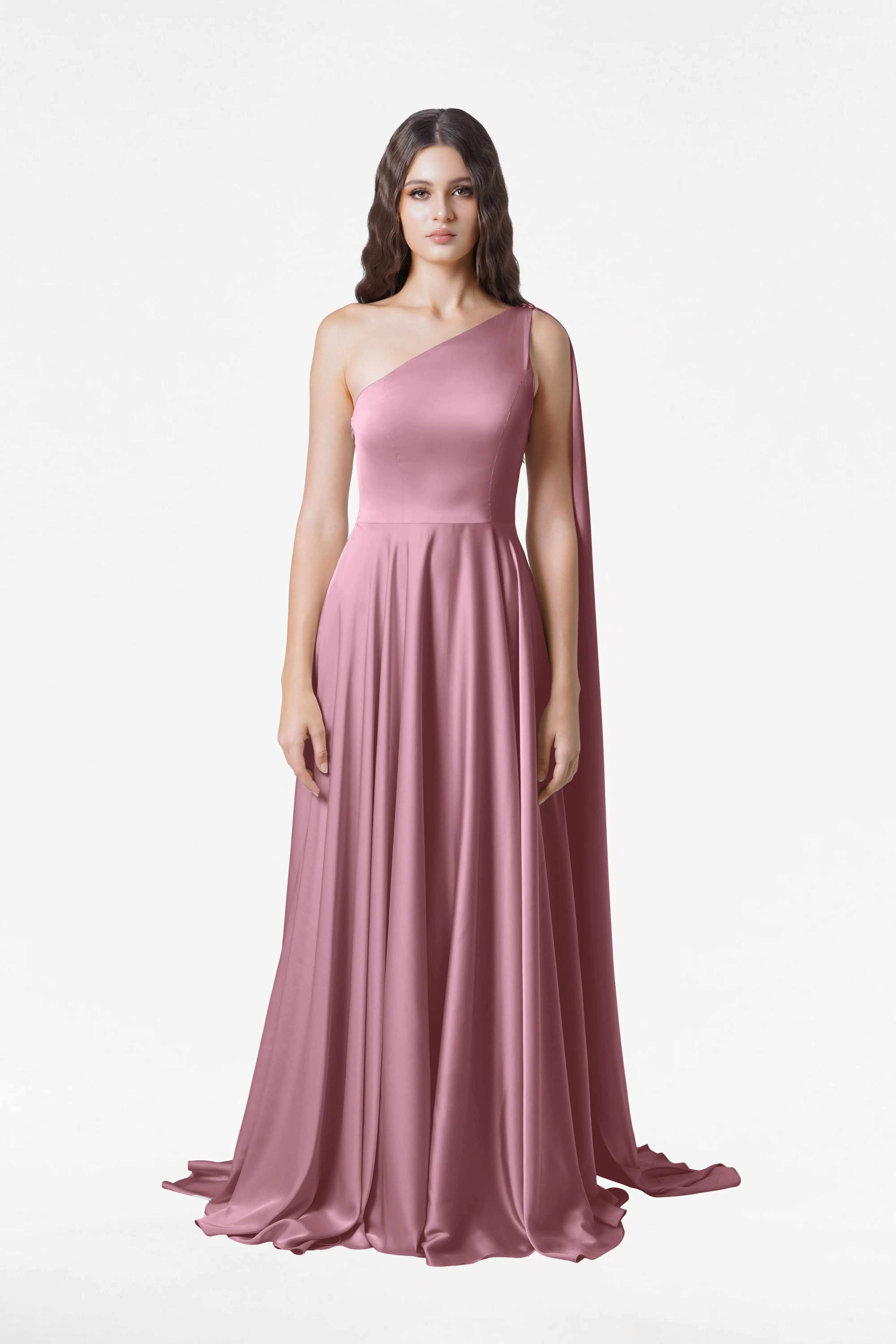A woman stands against a white background wearing the TH&TH Lyra Bridesmaid Dress in rose pink satin—a modern, size-inclusive gown with a flowing skirt and one-shoulder cape detail.