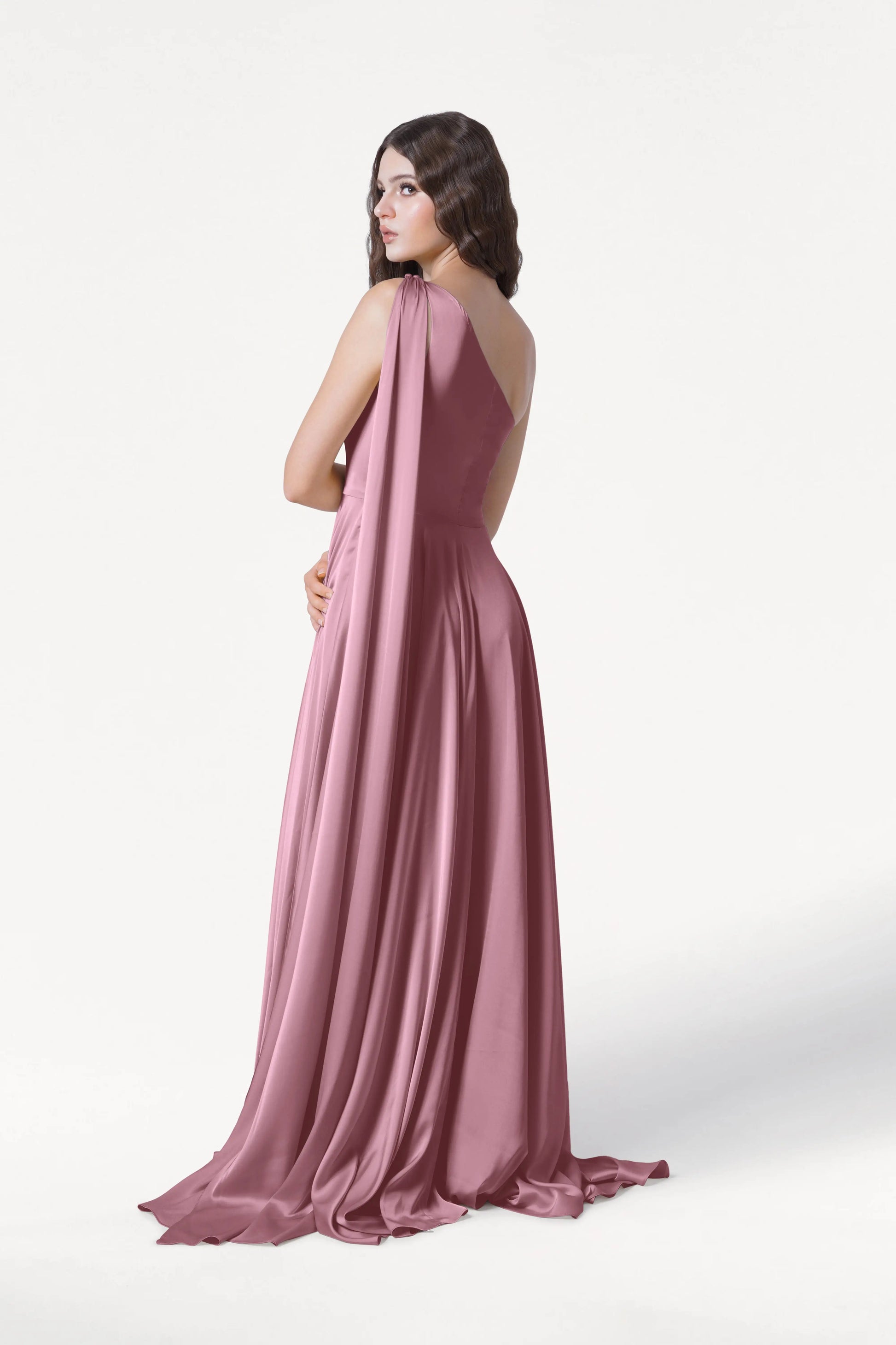 A woman with long, wavy brown hair models the TH&TH Lyra Bridesmaid Dress in rose pink satin, its elegant drape flowing as she poses partly turned against a plain white background.