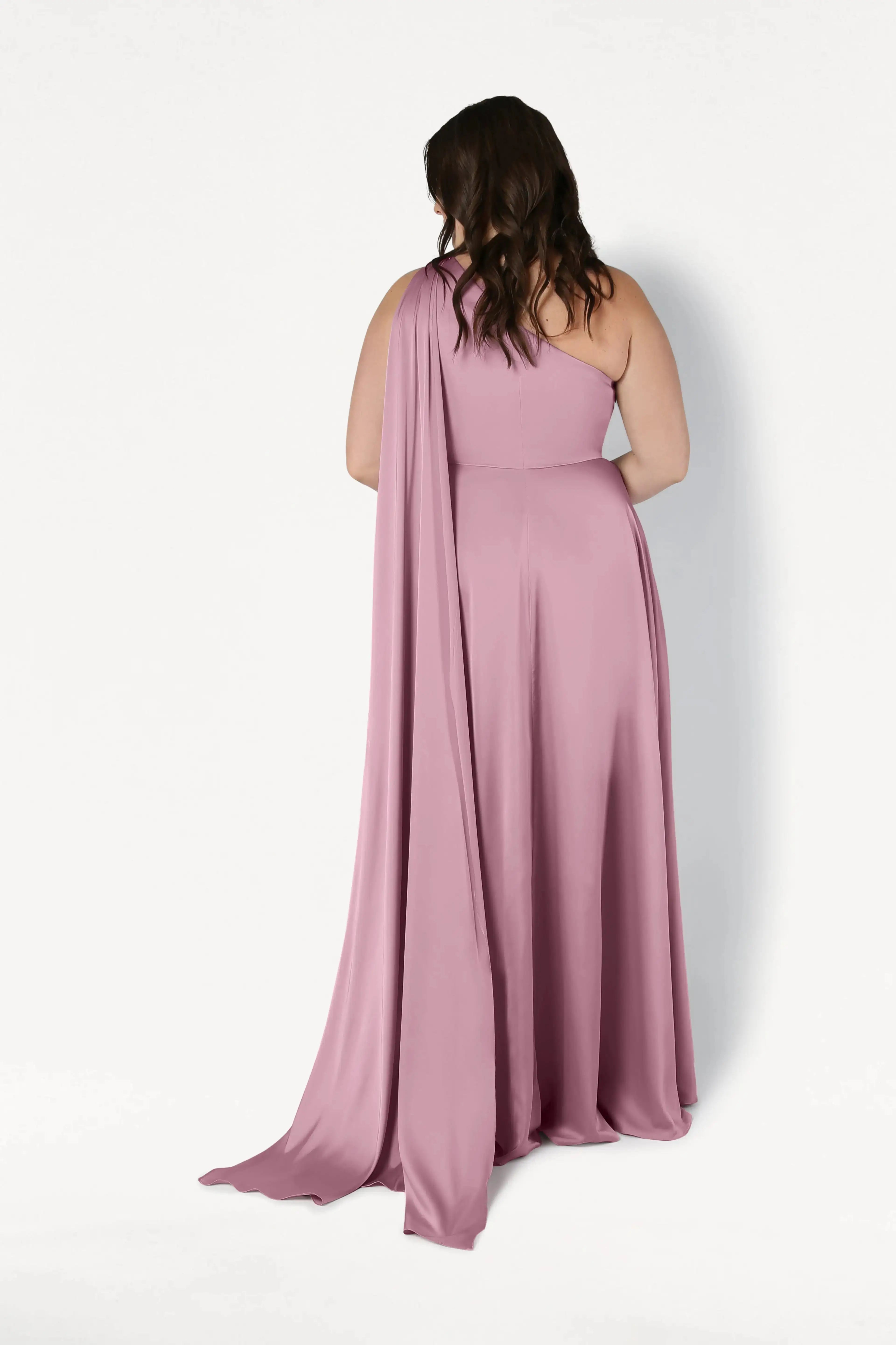 A woman with wavy brown hair stands facing away in the Lyra Bridesmaid Dress by TH&TH—a floor-length, rose pink satin one-shoulder gown with a draped sash—against a plain white background.