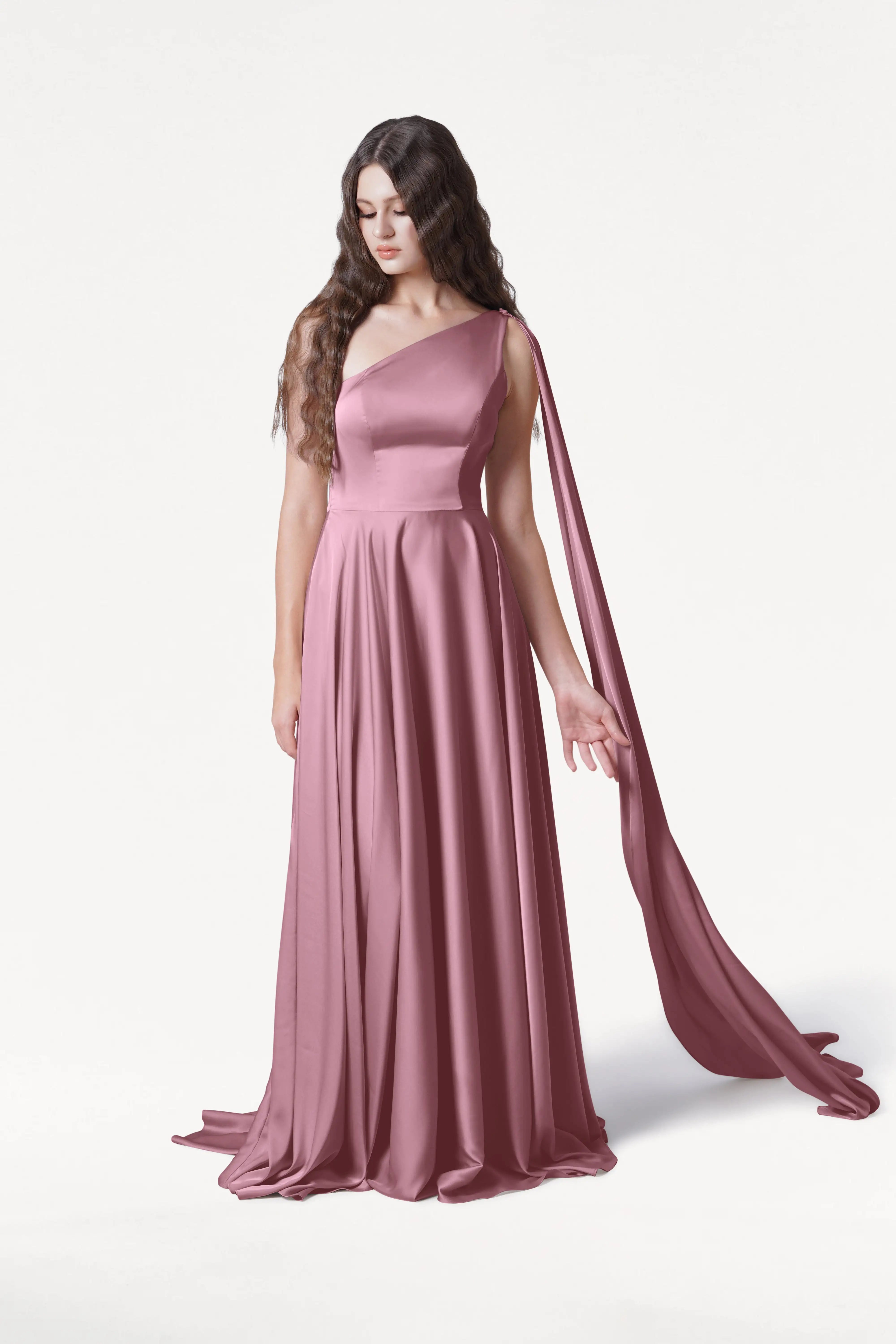 A woman with long wavy brown hair wears the Lyra Bridesmaid Dress in Rose Pink Satin by TH&TH, a size-inclusive, one-shoulder gown with a draped sash, standing against a plain white background and looking slightly downward.