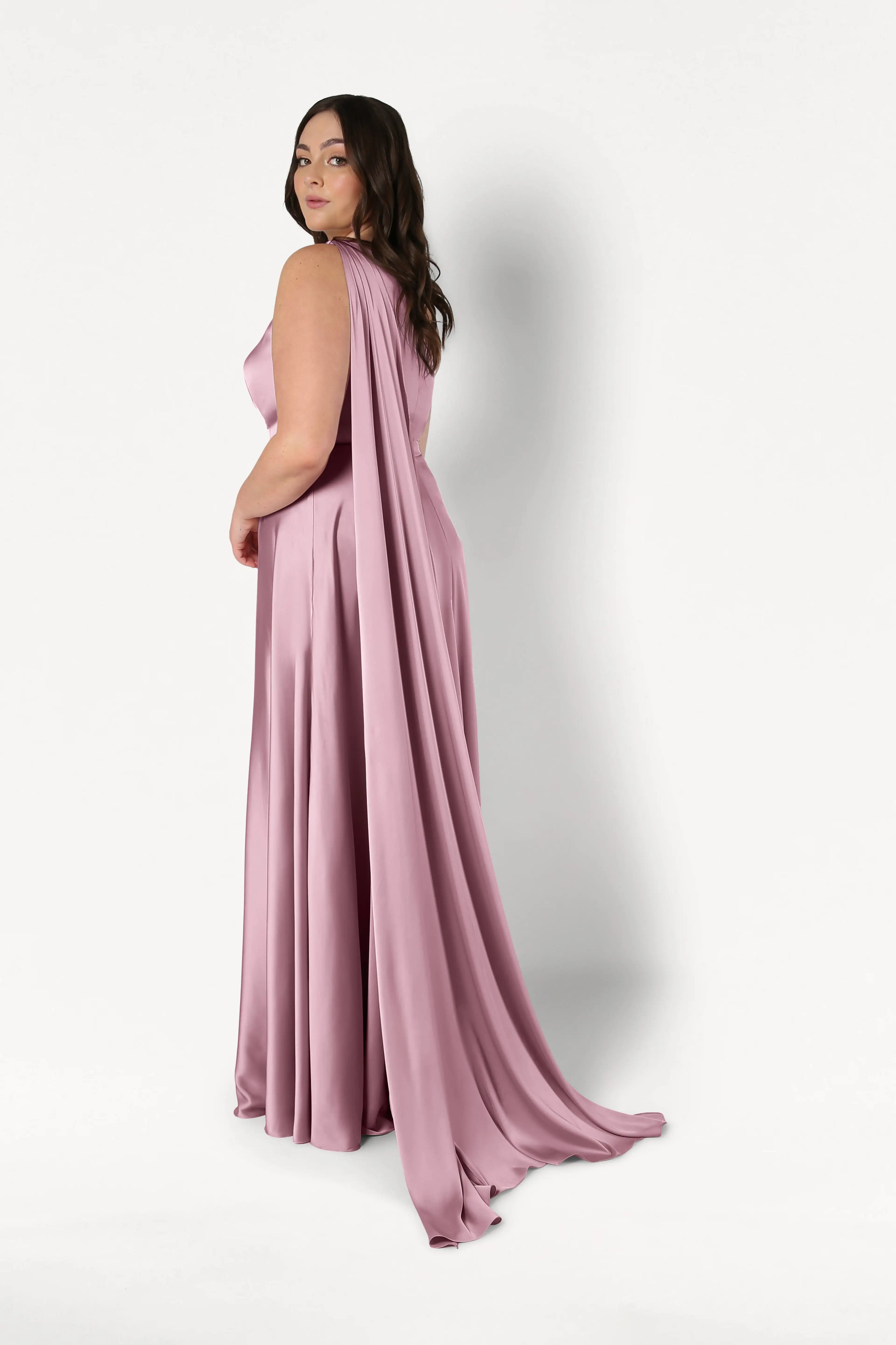 A woman models the TH&TH Lyra Bridesmaid Dress in rose pink satin, featuring a flowing, size-inclusive fit and long cape, as she poses against a plain white background, glancing over her shoulder at the camera.