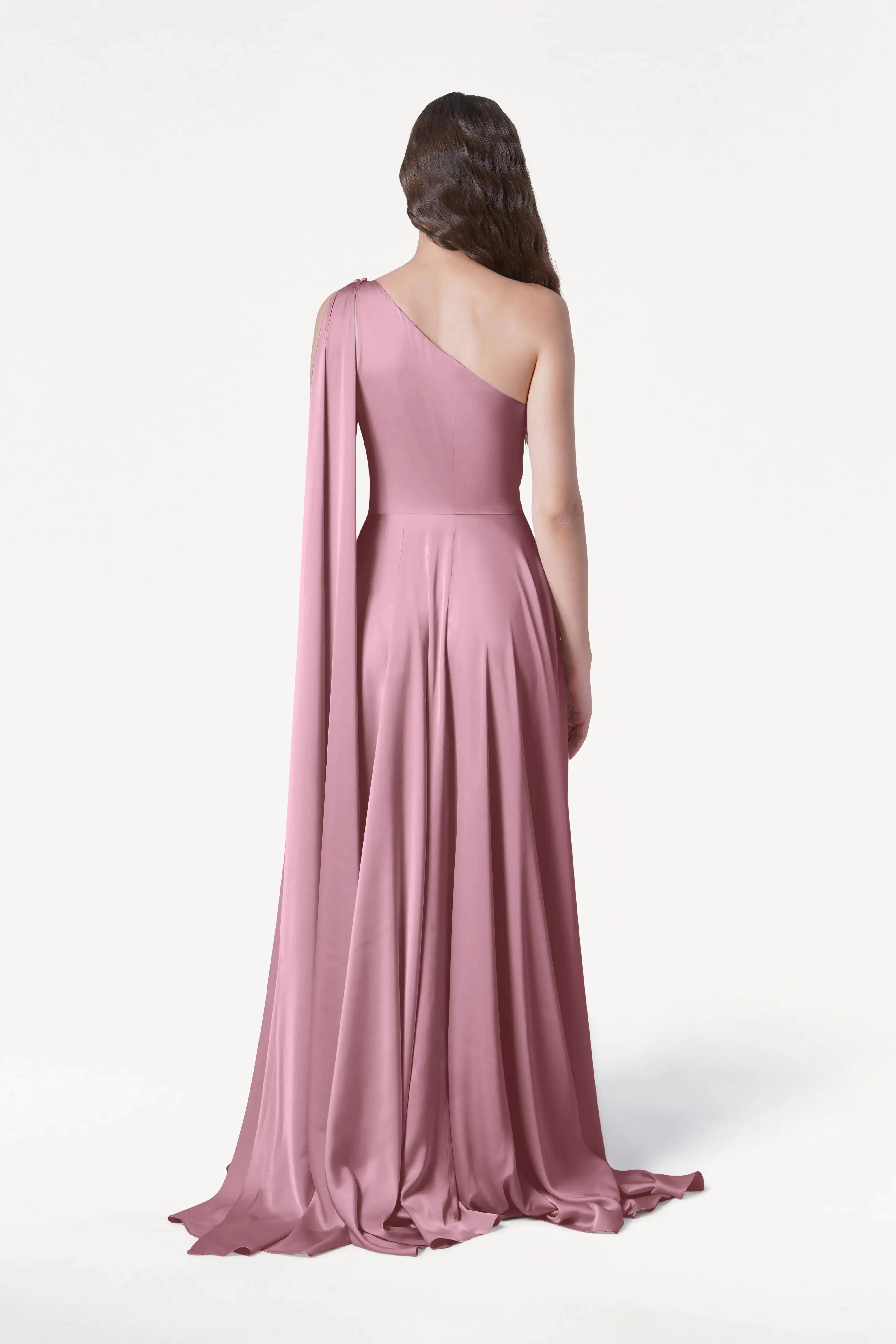 A woman with wavy hair faces away, wearing the TH&TH Lyra Bridesmaid Dress in rose pink satin, featuring a modern one-shoulder cut and draped cape detail, set against a plain white background.
