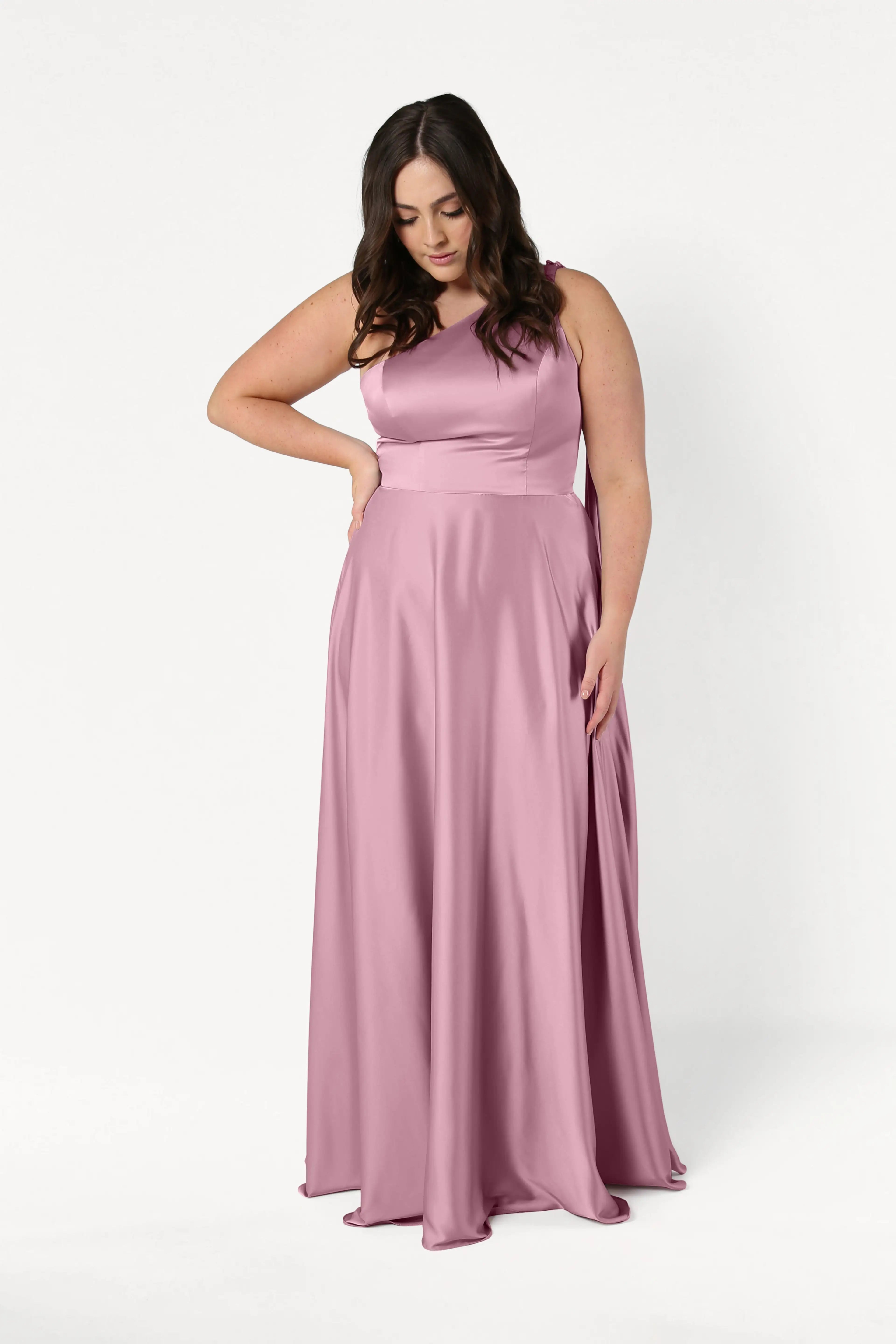 A woman models the TH&TH Lyra Bridesmaid Dress in Rose Pink Satin, standing against a white background with one hand on her hip and the other at her side, looking down.