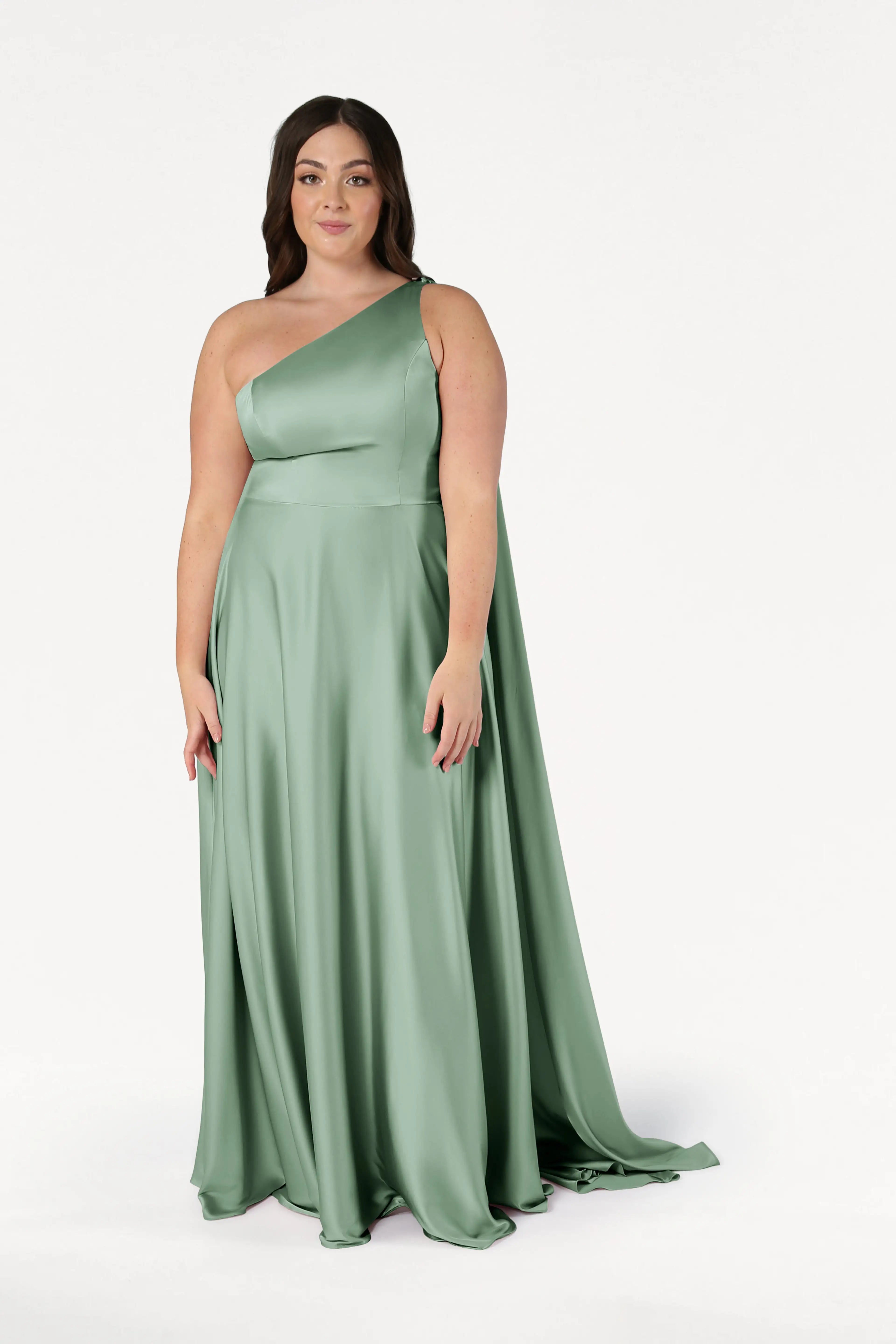A woman in the TH&TH Lyra Bridesmaid Dress in Sage Green Satin poses against a white background. The size-inclusive gown features a one-shoulder design, fitted bodice, and flowing floor-length skirt.