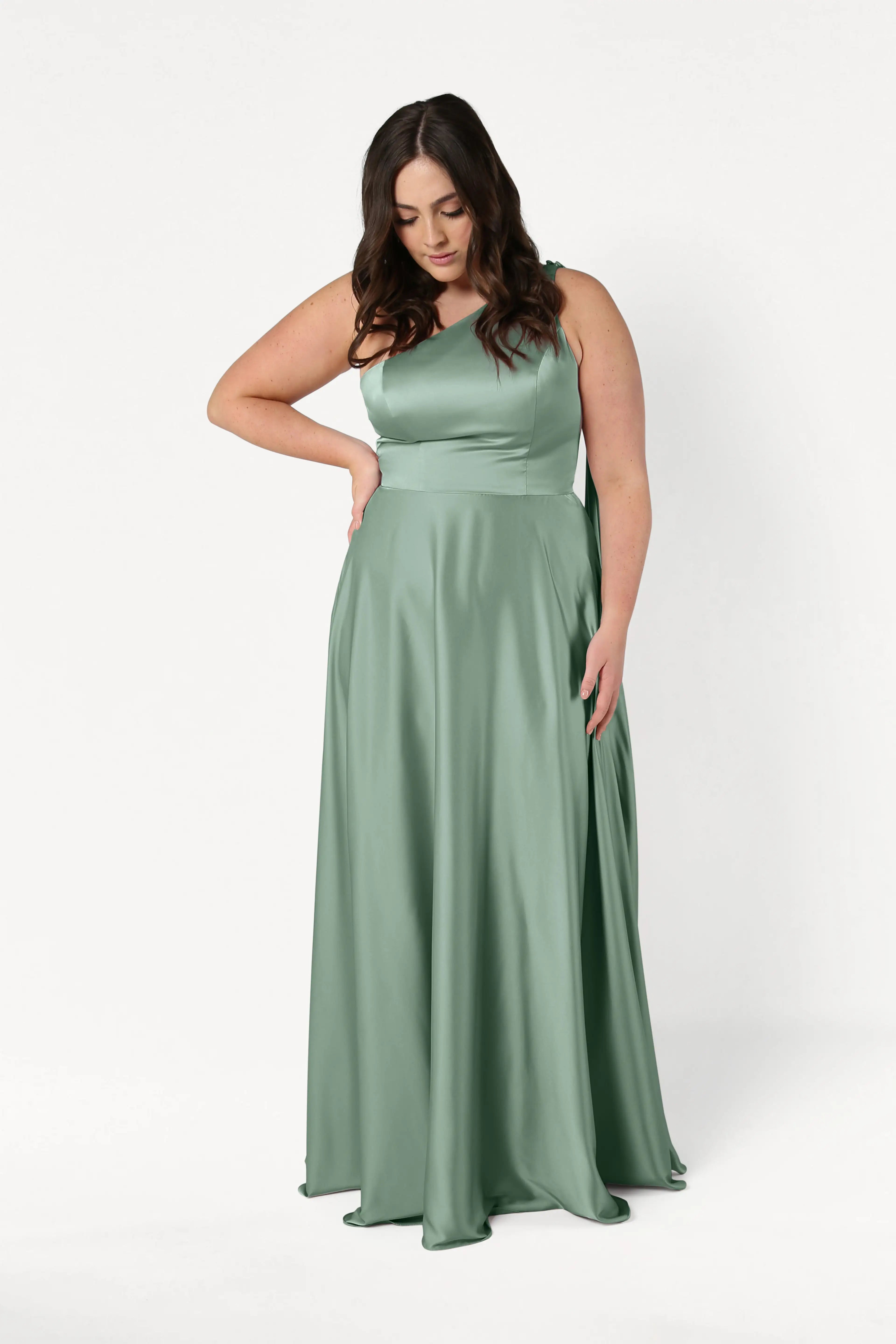 A woman models the TH&TH Lyra Bridesmaid Dress in sage green satin, a size-inclusive, one-shoulder style, standing against a white background with one hand on her waist and the other holding her dress.