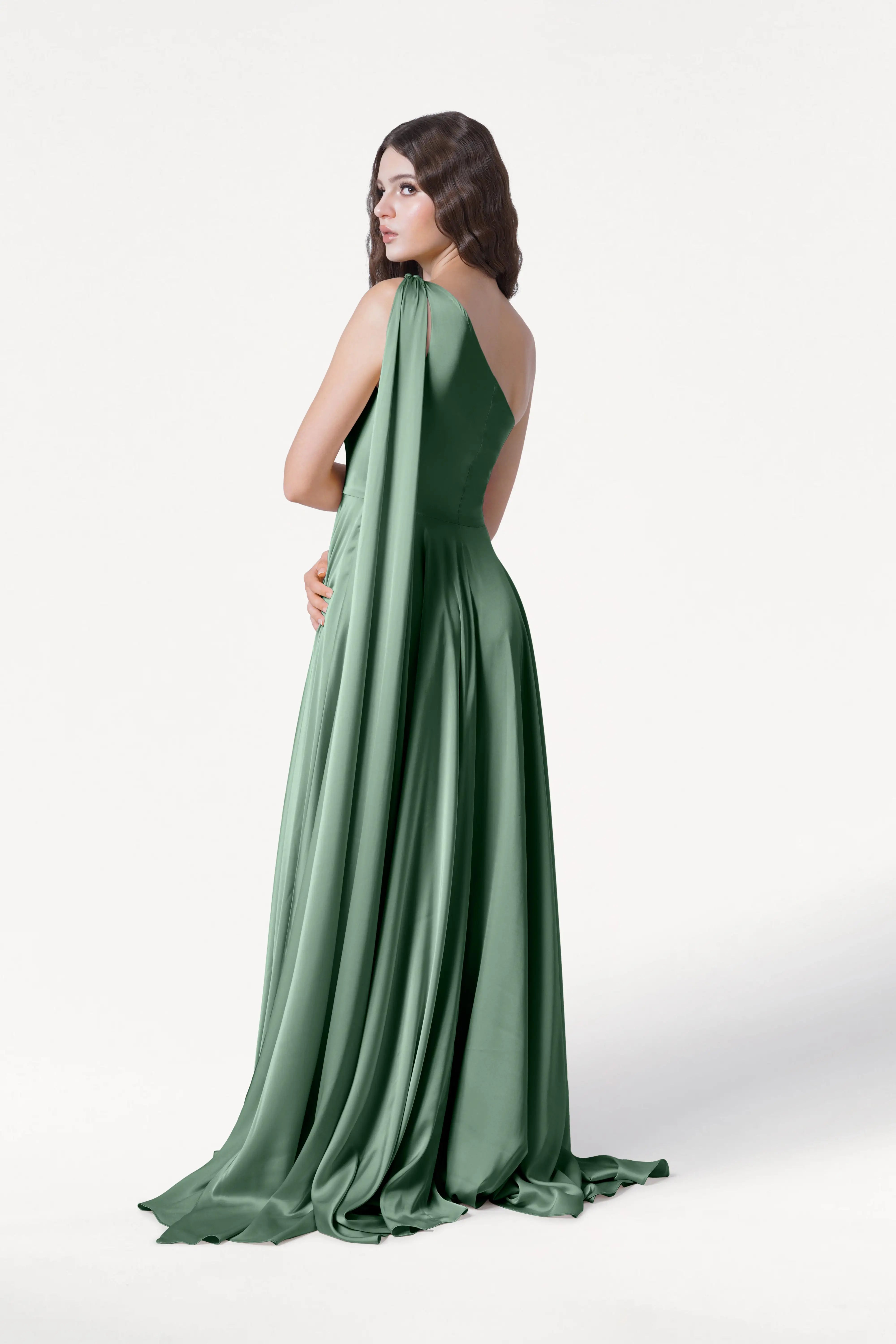 A woman poses against a white background in the TH&TH Lyra Bridesmaid Dress, featuring one-shoulder styling and flowing sage green satin.