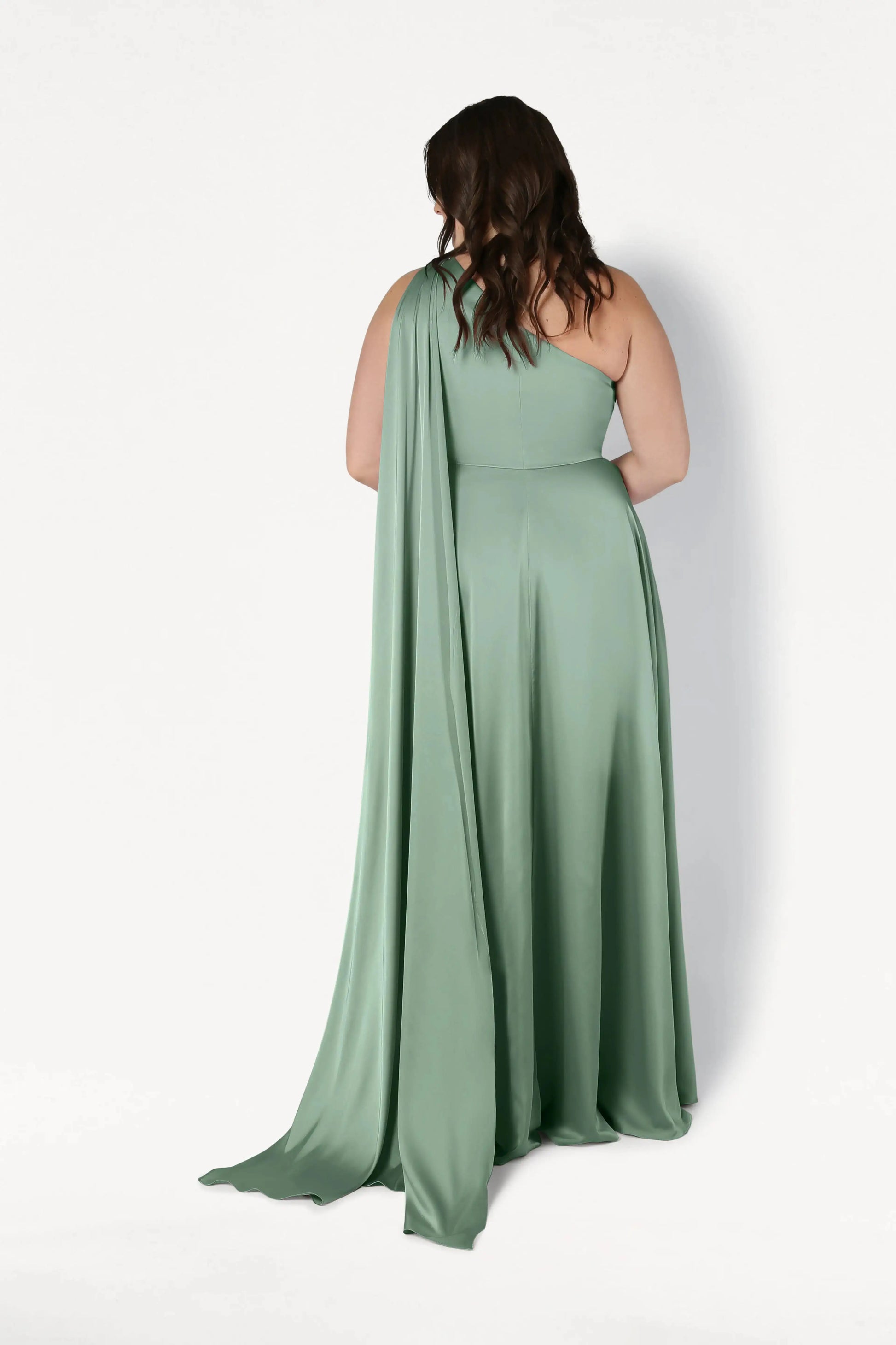 A woman with long, wavy brown hair models the TH&TH Lyra Bridesmaid Dress in sage green satin, a size-inclusive one-shoulder gown with a flowing drape. She stands against a white background, facing away from the camera.