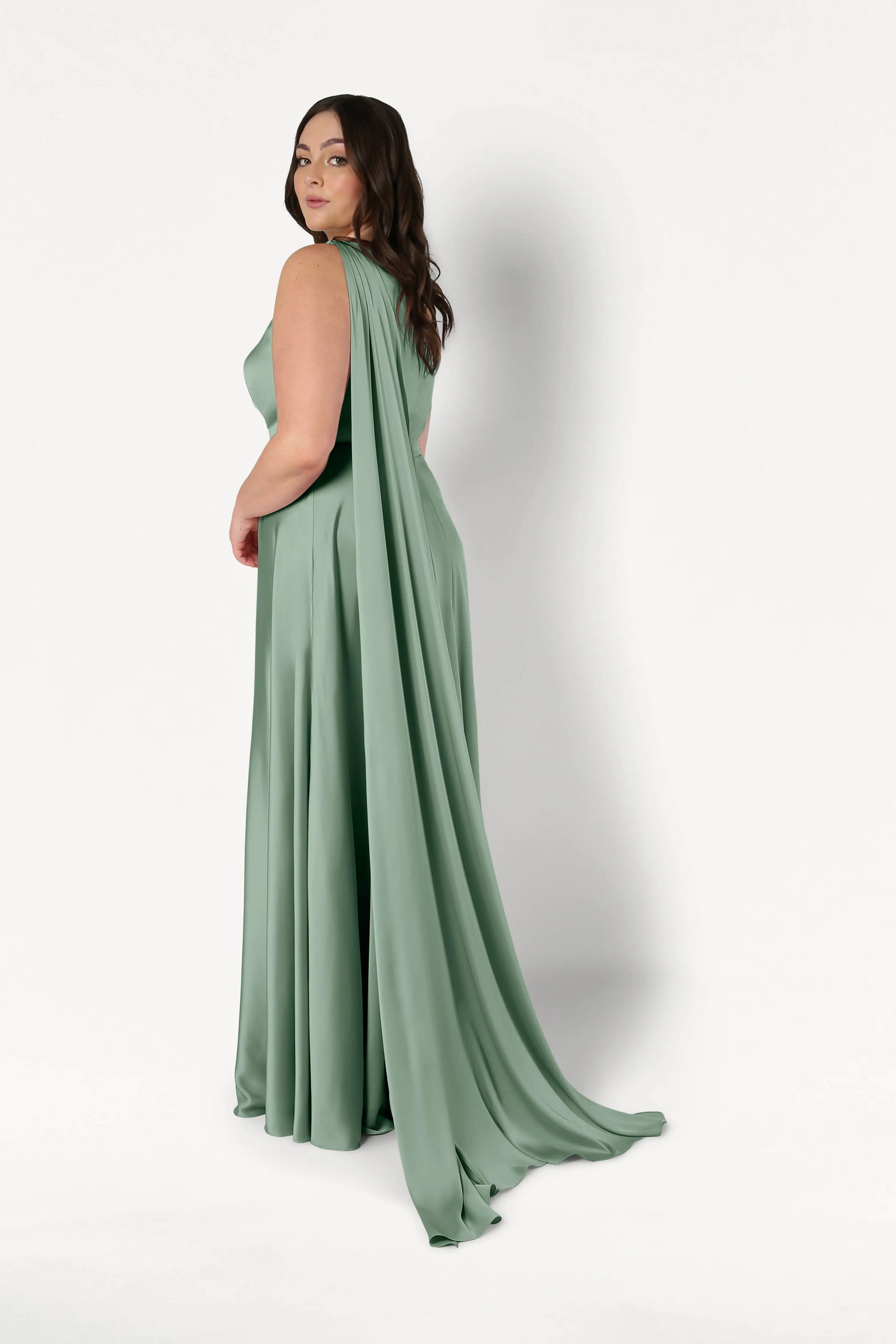 A woman models the TH&TH Lyra Bridesmaid Dress in sage green satin. The size-inclusive, one-shoulder gown features a dramatic cape-like train and is set against a white background as she looks over her shoulder.