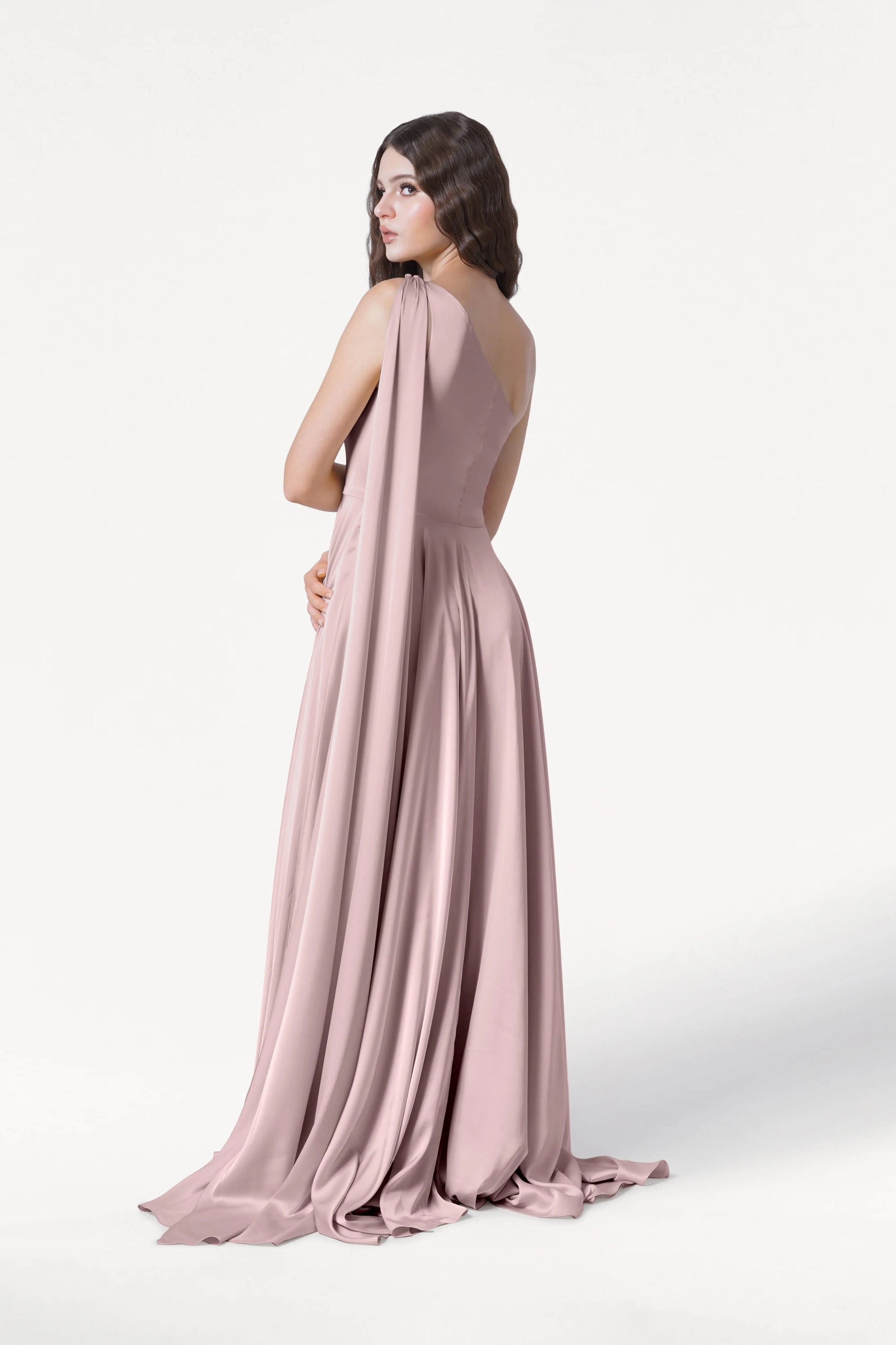 A woman with long wavy brown hair models the TH&TH Lyra Bridesmaid Dress in Smoked Blush Satin—a size-inclusive, floor-length gown with a flowing train and draped fabric—standing against a plain white background, looking over her shoulder.