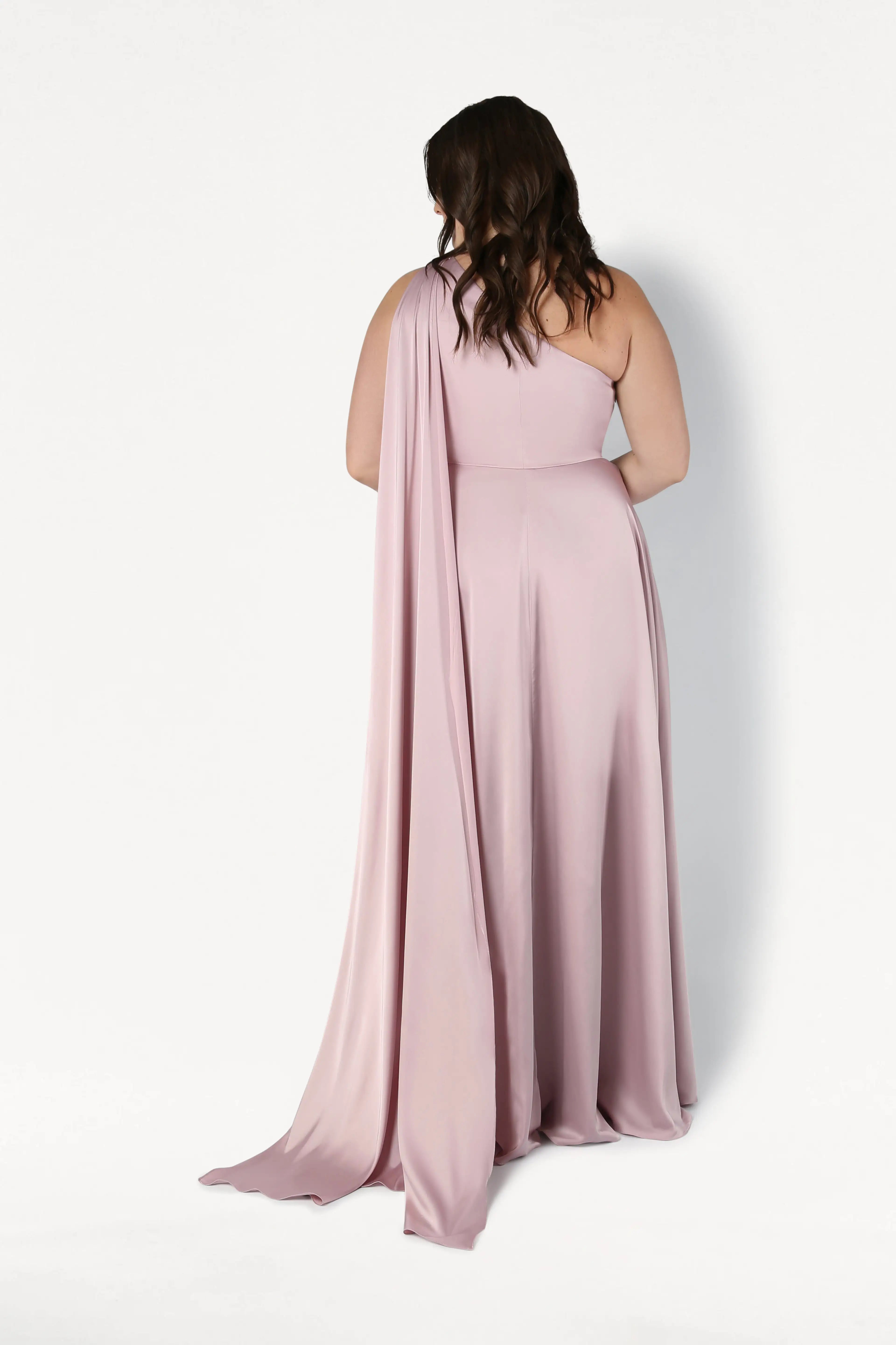 A woman with wavy brown hair stands with her back to the camera, wearing the size-inclusive Lyra Bridesmaid Dress in Smoked Blush Satin by TH&TH. The one-shoulder satin gown flows elegantly, featuring draped fabric against a plain white background.