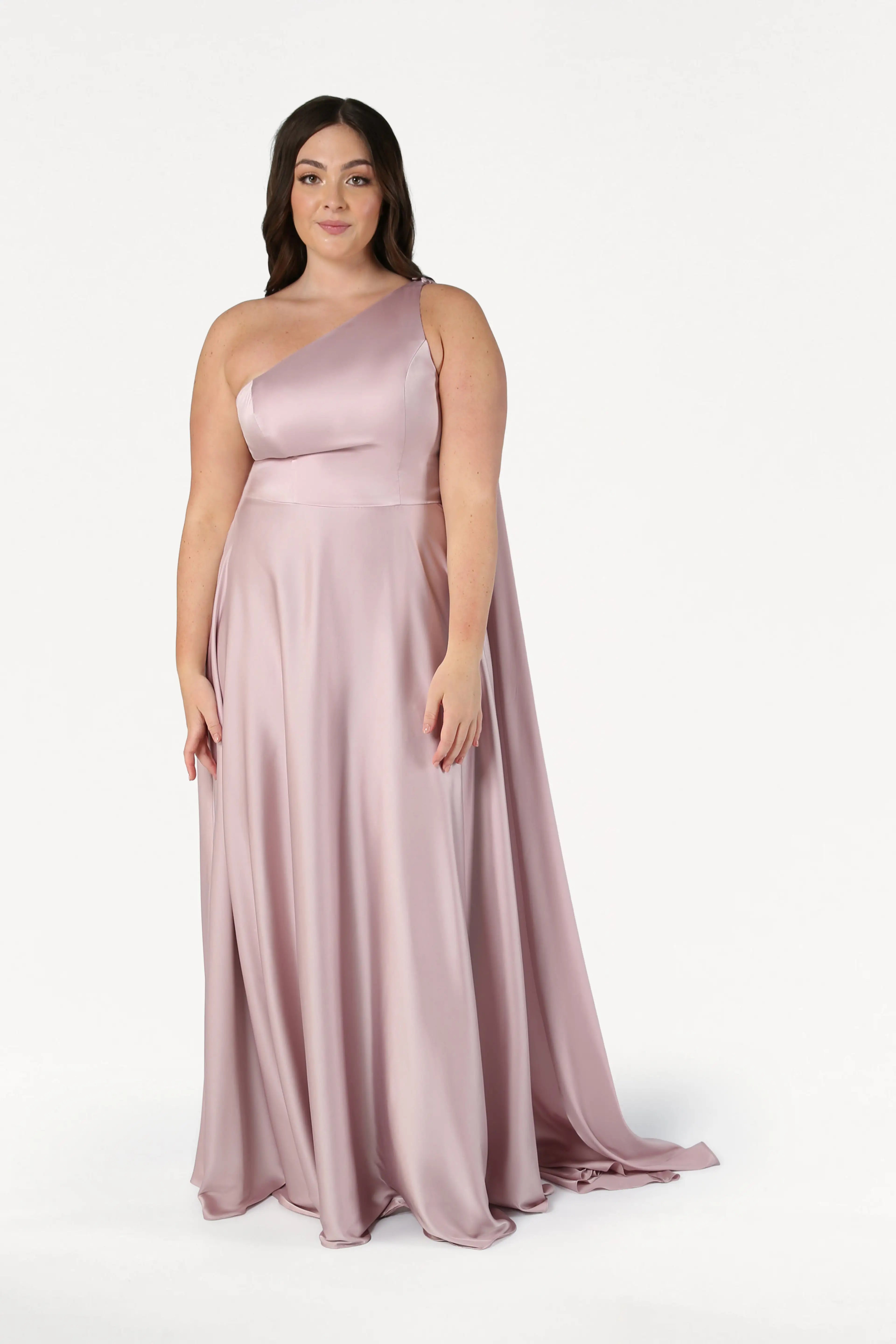 A woman in the Lyra Bridesmaid Dress in Smoked Blush Satin by TH&TH poses against a white background, softly smiling. This elegant, size-inclusive one-shoulder gown is perfect for every celebration.