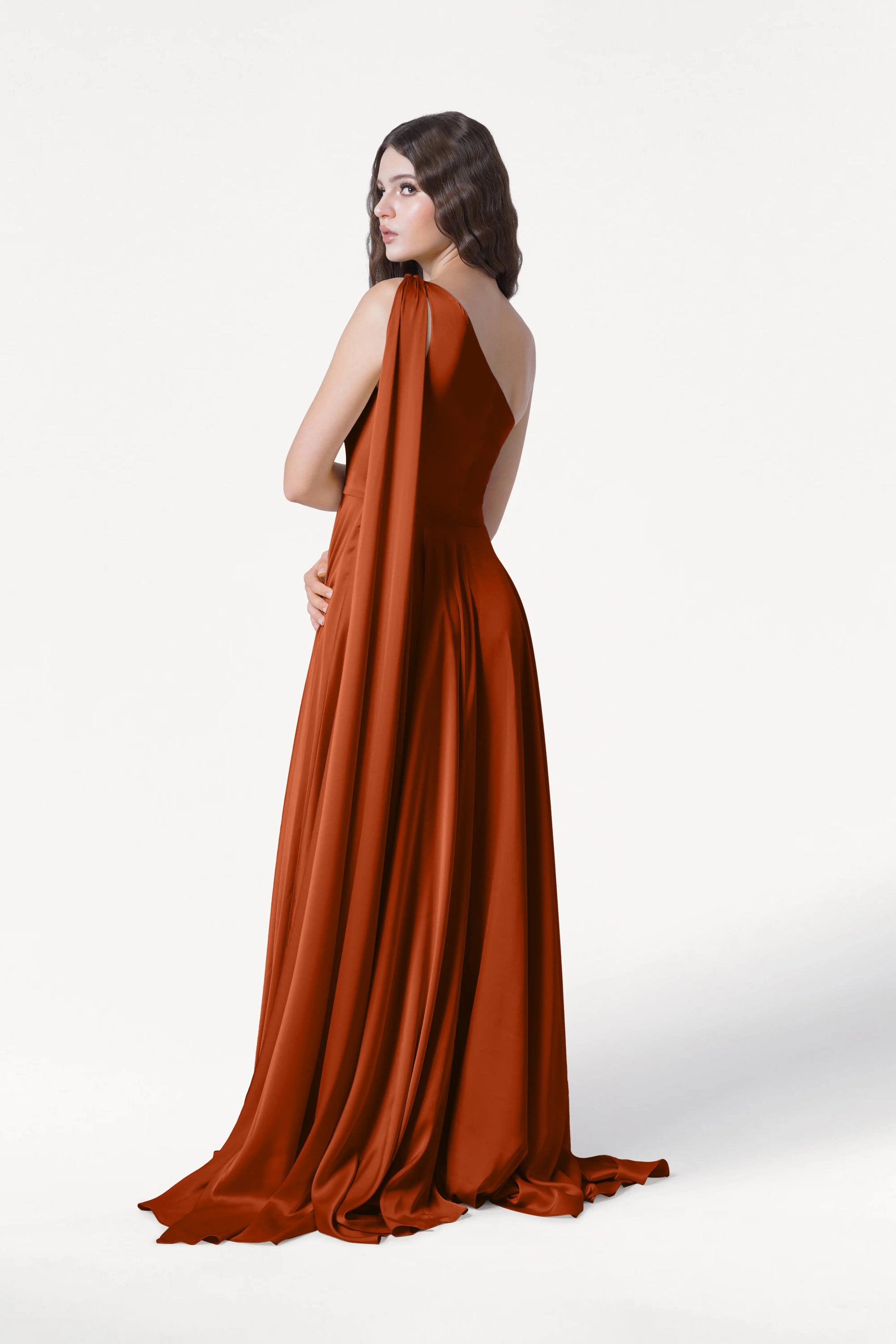 A woman with wavy brown hair models the TH&TH Lyra Bridesmaid Dress in Terracotta Satin—a floor-length, one-shoulder gown with a long draped train—standing slightly turned against a plain white background.