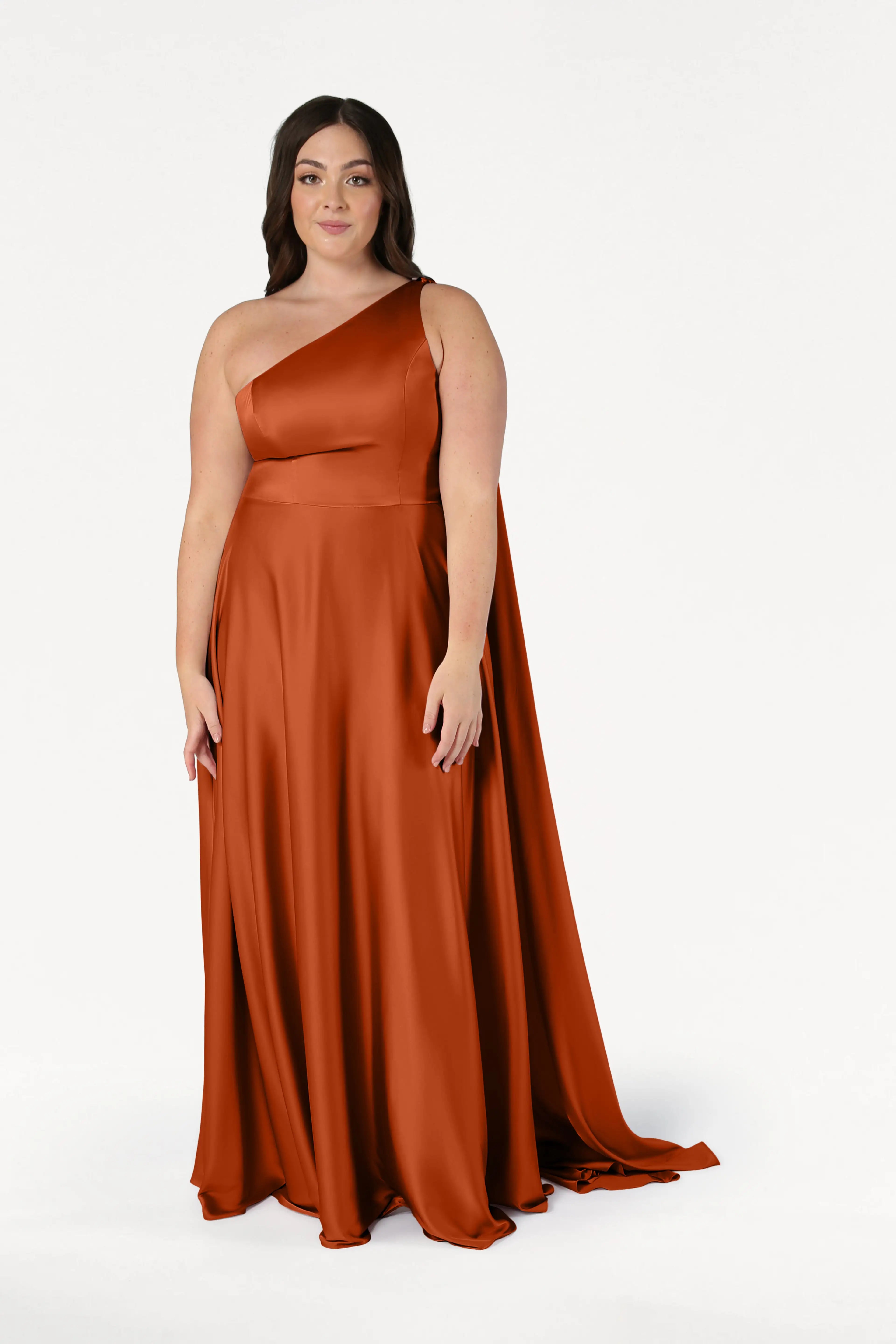 A woman stands smiling in the TH&TH Lyra Bridesmaid Dress in Terracotta Satin—a size-inclusive, floor-length, one-shoulder gown with a flowing train—set against a plain white background.