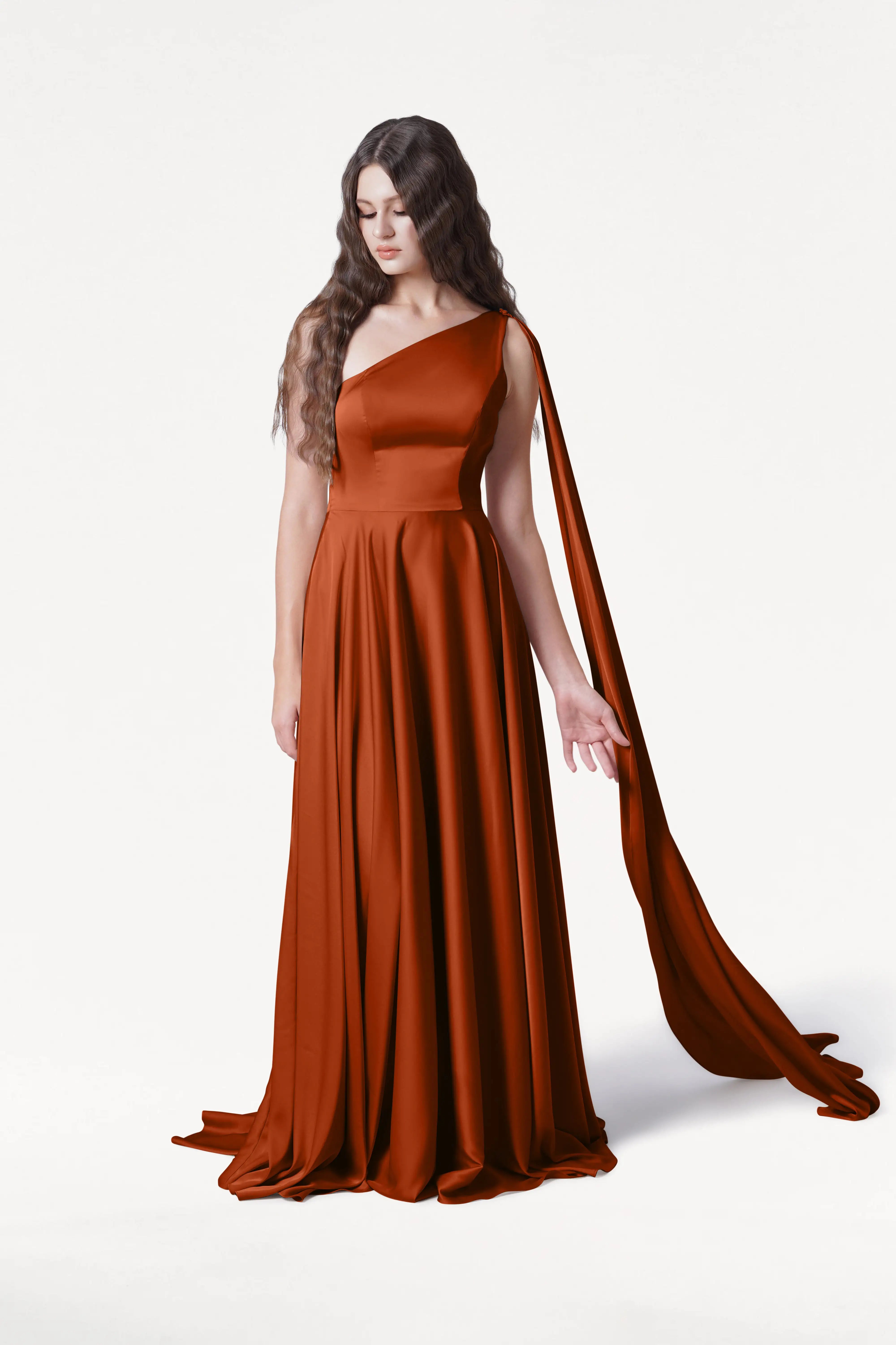 A woman with long, wavy brown hair models the TH&TH Lyra Bridesmaid Dress in Terracotta Satin—a modern, one-shoulder gown with a draped sash—standing against a white background and gazing slightly downward.