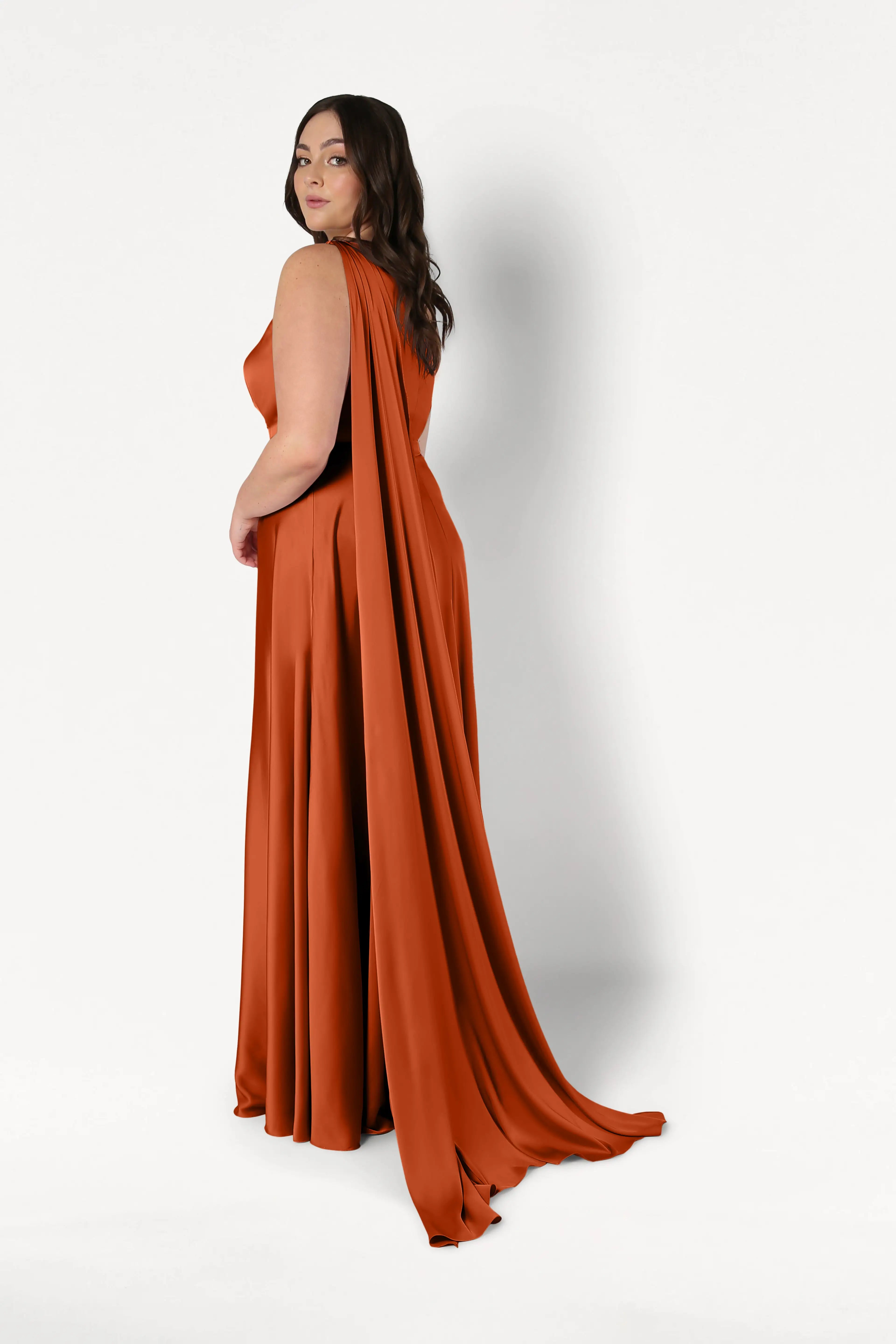 A woman with long brown hair models the size-inclusive Lyra Bridesmaid Dress in Terracotta Satin by TH&TH, a floor-length gown with a dramatic draped cape. She stands turned to the side, looking over her shoulder against a plain white background.