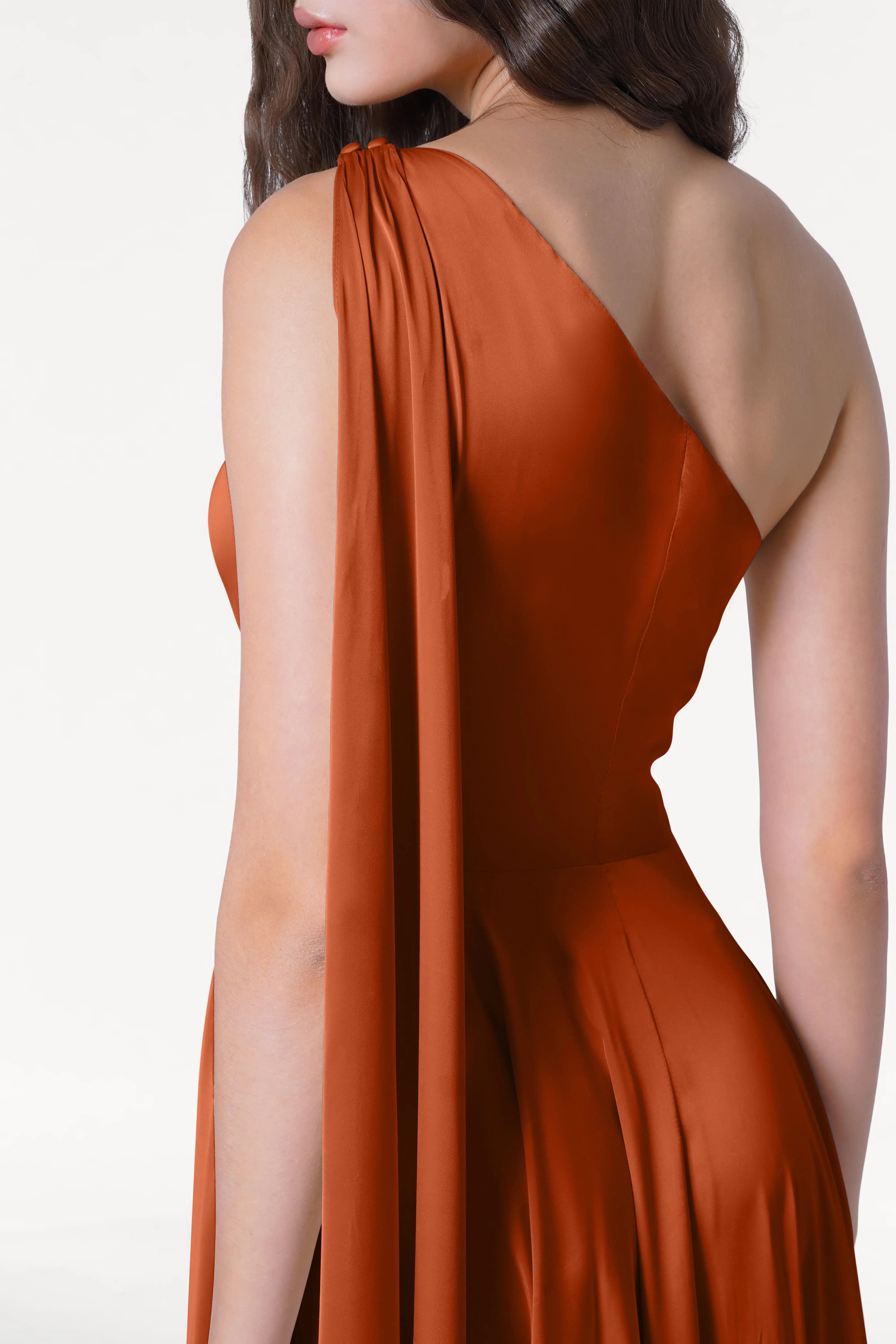 A person with long, wavy brown hair models the size-inclusive Lyra Bridesmaid Dress by TH&TH in terracotta satin with a one-shoulder drape, standing against a plain white background.