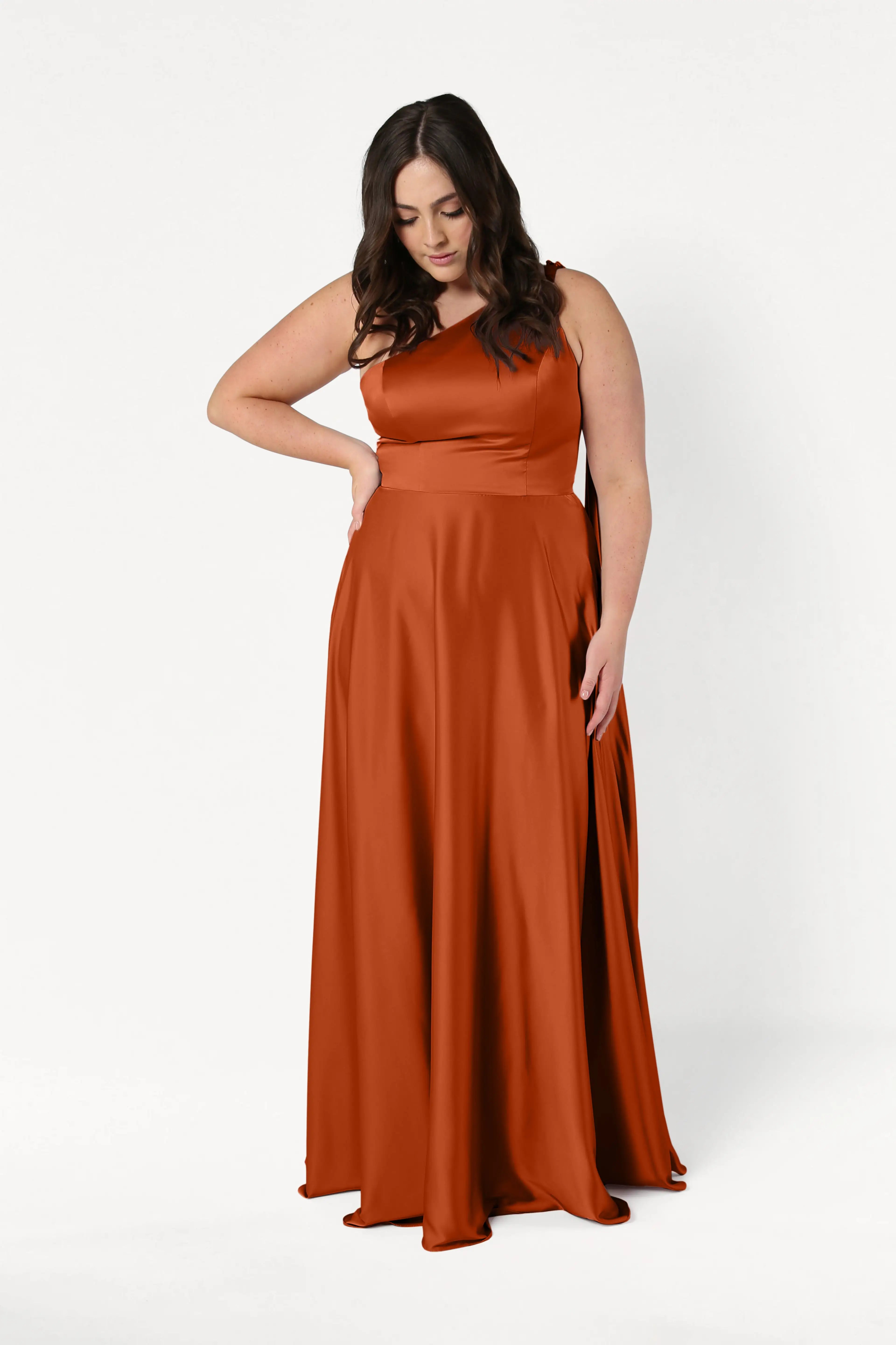 A woman with long dark hair models the Lyra Bridesmaid Dress in Terracotta Satin by TH&TH. This size-inclusive, sleeveless, floor-length gown is shown against a plain white background as she poses with one hand on her hip.