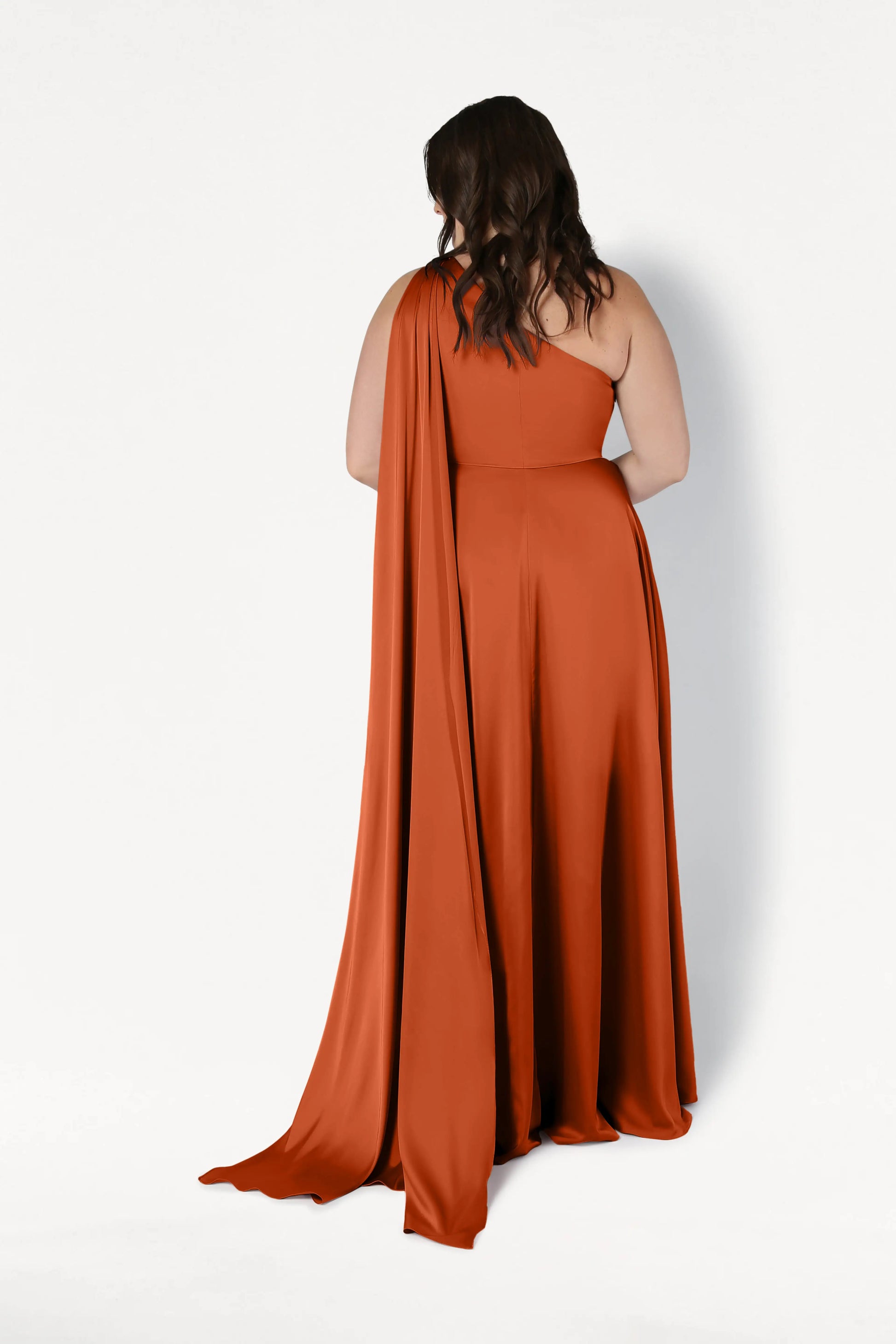 A woman with wavy dark hair faces away, wearing the TH&TH Lyra Bridesmaid Dress in Terracotta Satin—a flowing, size-inclusive one-shoulder gown with a long draped train—against a plain white background.