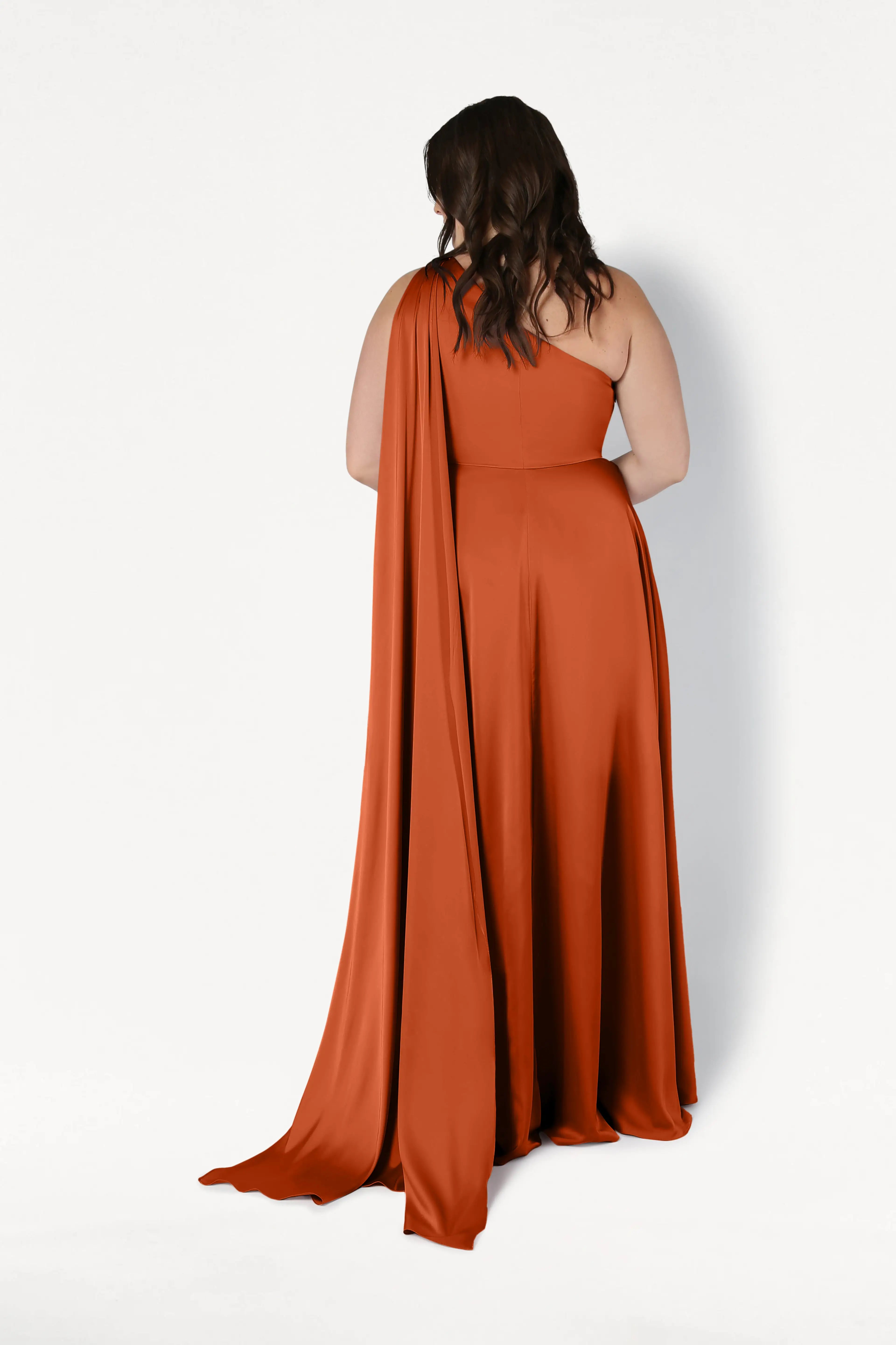 A woman with wavy dark hair faces away, wearing the TH&TH Lyra Bridesmaid Dress in Terracotta Satin—a flowing, size-inclusive one-shoulder gown with a long draped train—against a plain white background.