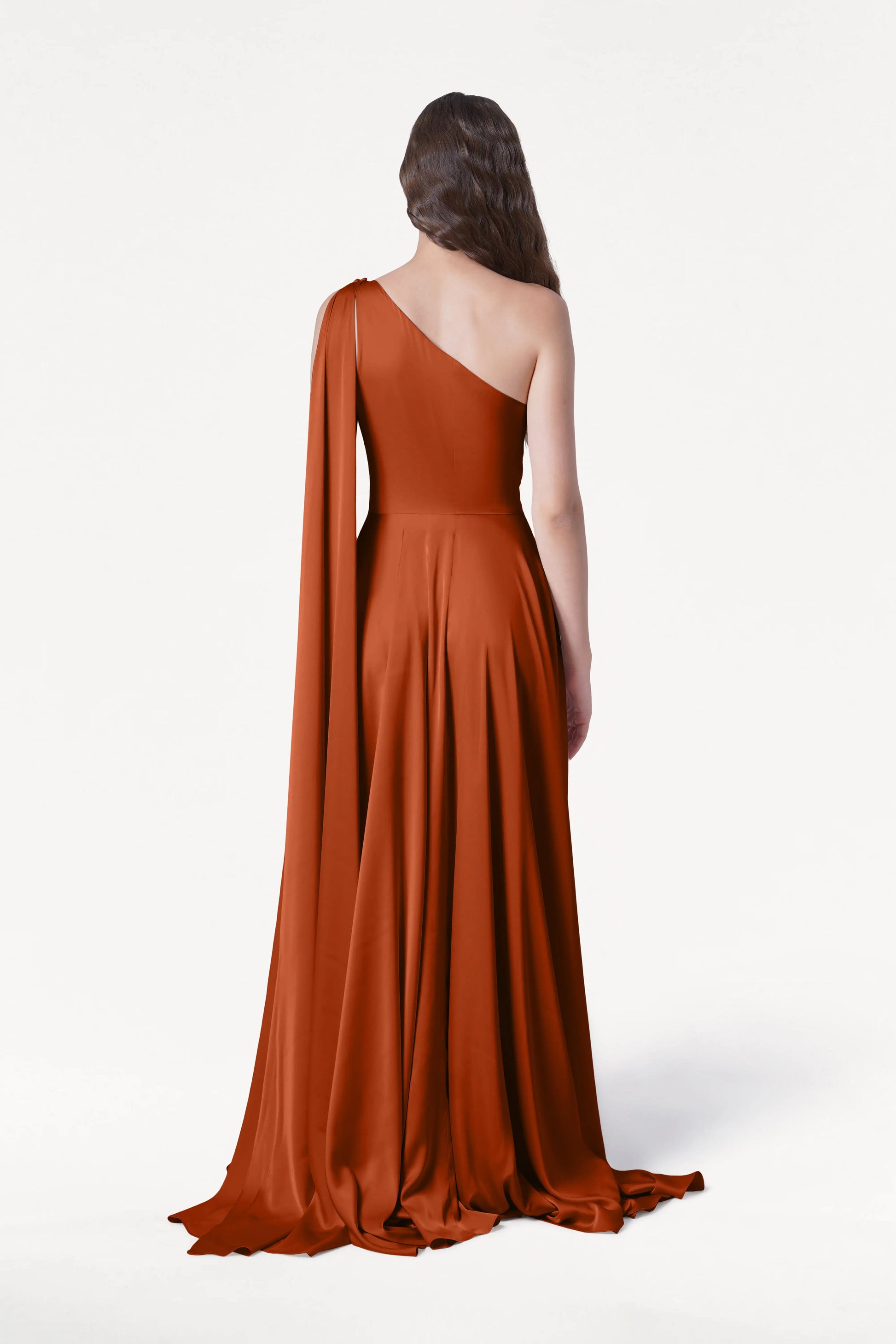 A woman faces away in the Lyra Bridesmaid Dress by TH&TH, a floor-length, terracotta satin one-shoulder gown with a draped sleeve and flowing fabric. Her brown hair is styled in loose waves against a plain white background.