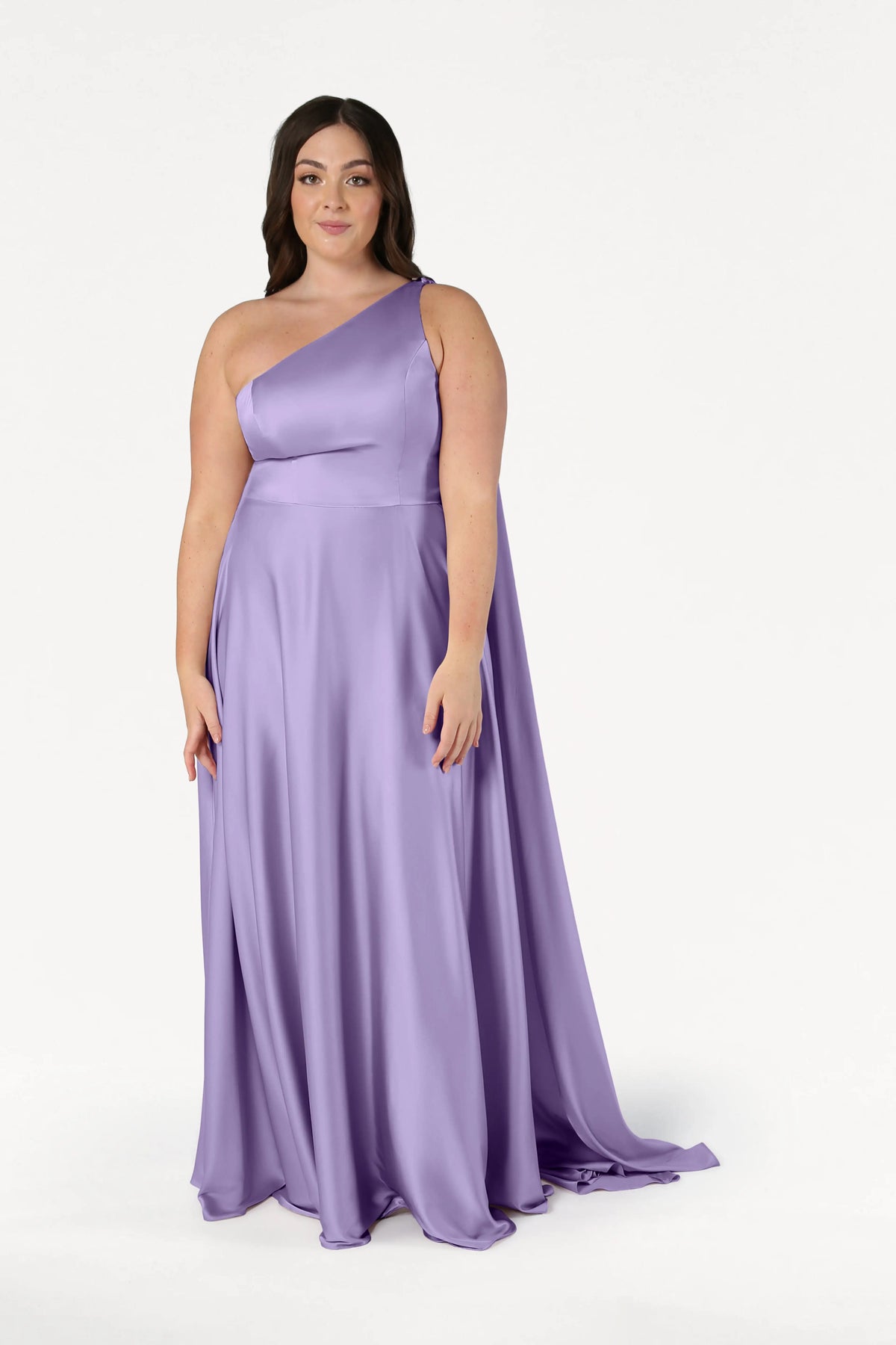 A woman wears the size-inclusive Lyra Bridesmaid Dress in Violet Satin by TH&TH—a floor-length, one-shoulder gown with a flowing skirt—standing against a plain white background.