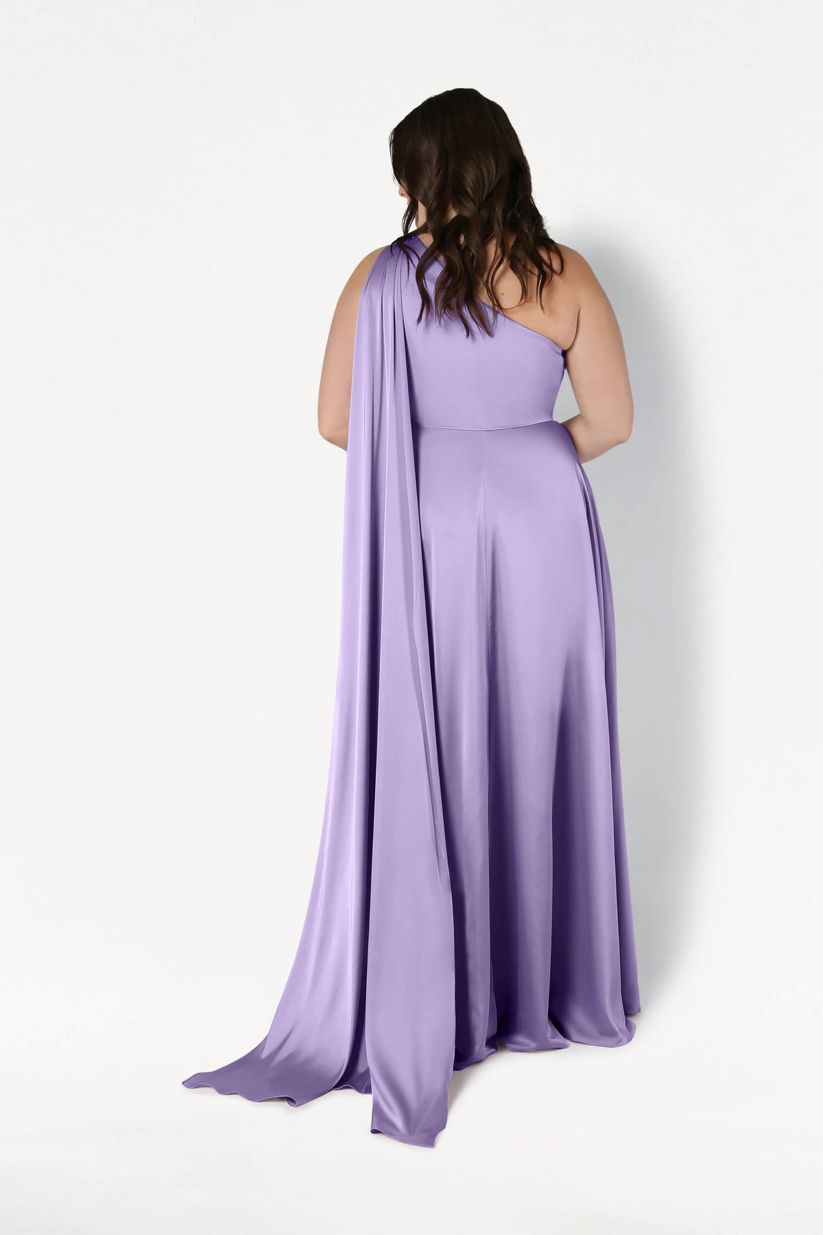 A woman with wavy brown hair models the size-inclusive Lyra Bridesmaid Dress in Violet Satin by TH&TH, featuring a flowing one-shoulder design with a draped cape. She stands with her back to the camera against a white background, highlighting its elegant silhouette.