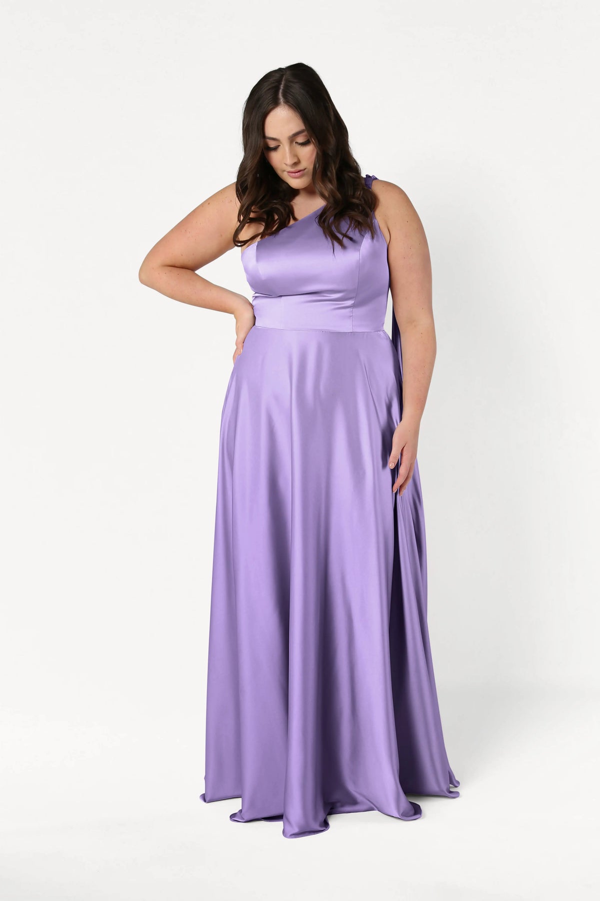 A woman models the size-inclusive TH&TH Lyra Bridesmaid Dress in Violet Satin, floor-length and elegant, standing against a plain white background with one hand on her hip and the other holding her dress hem.