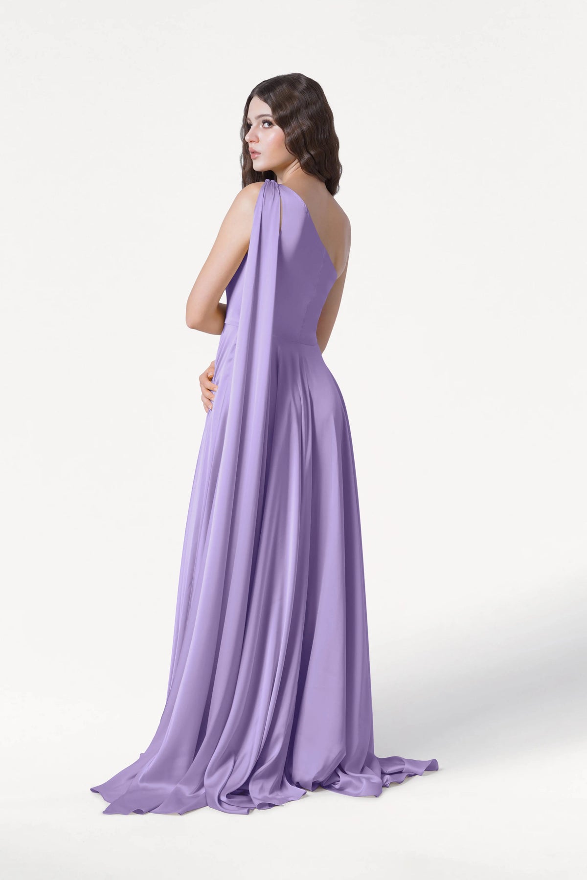 A woman with long brown hair models the Lyra Bridesmaid Dress in Violet Satin by TH&TH—a size-inclusive, one-shoulder gown—posing against a plain white background.