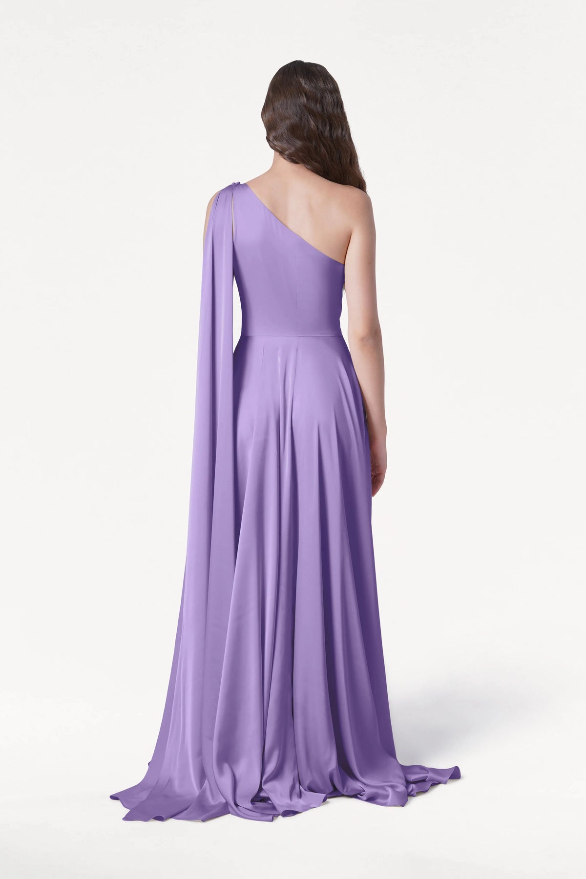 A woman with wavy brown hair faces away, wearing the size-inclusive Lyra Bridesmaid Dress in Violet Satin by TH&TH—a one-shoulder, floor-length gown with a draped, flowing train—set against a plain white background.