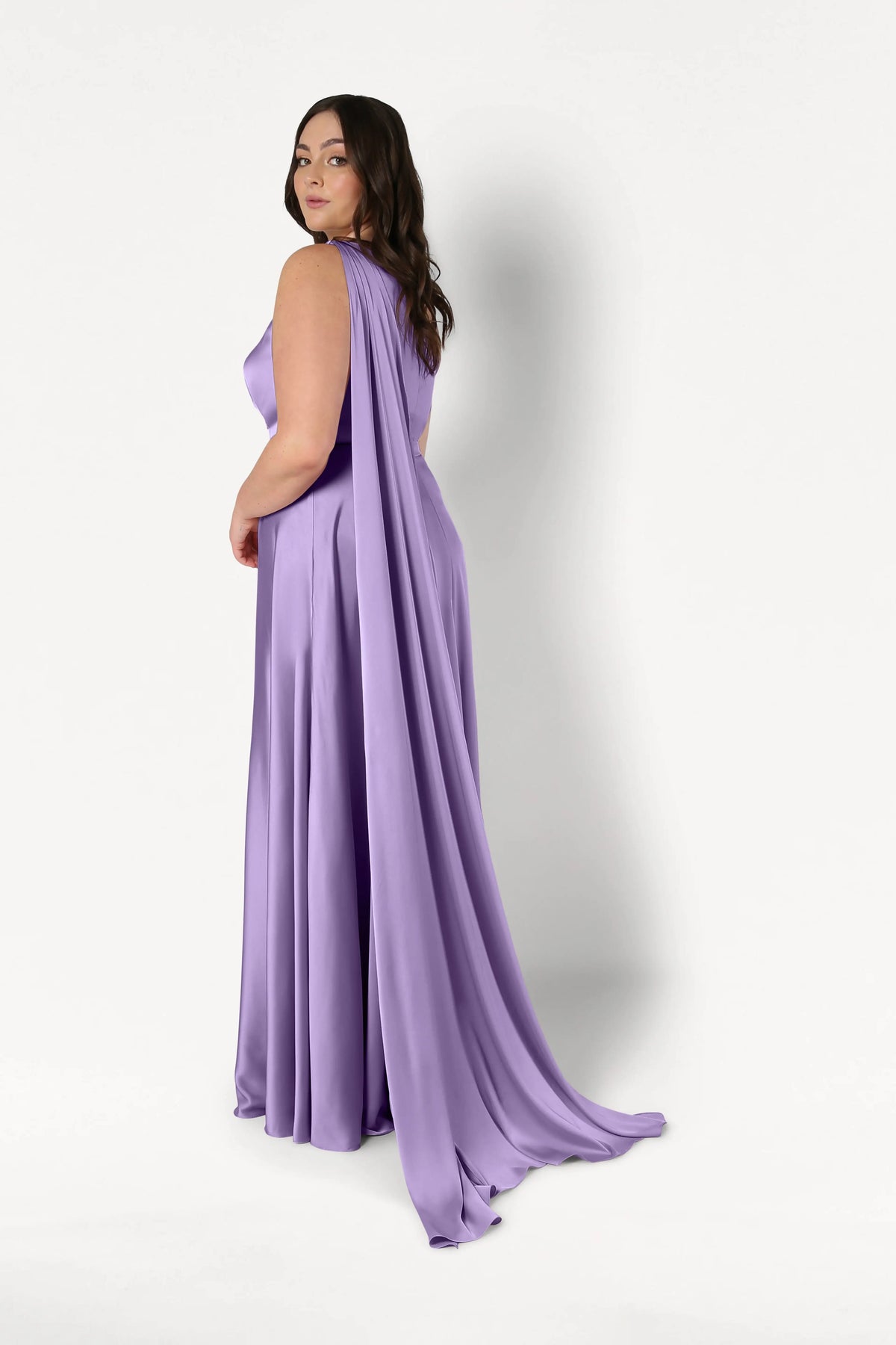 A woman poses against a white background in the TH&TH Lyra Bridesmaid Dress, featuring violet satin, inclusive sizing, and a cape-like train, as she looks over her shoulder.