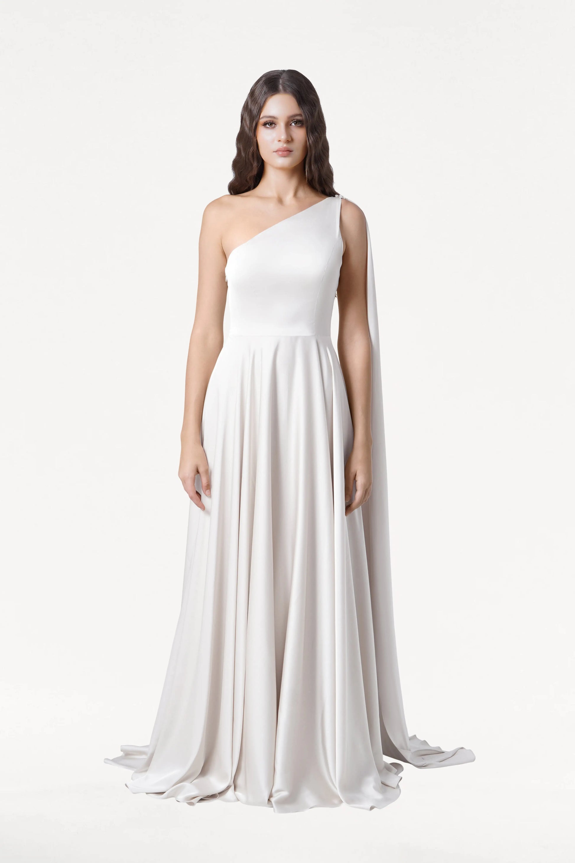 A woman with long, wavy dark hair and a neutral expression models the Lyra Dress in Ivory Satin by TH&TH—a size-inclusive, flowing, one-shoulder gown with a cape-like drape—against a plain white background.