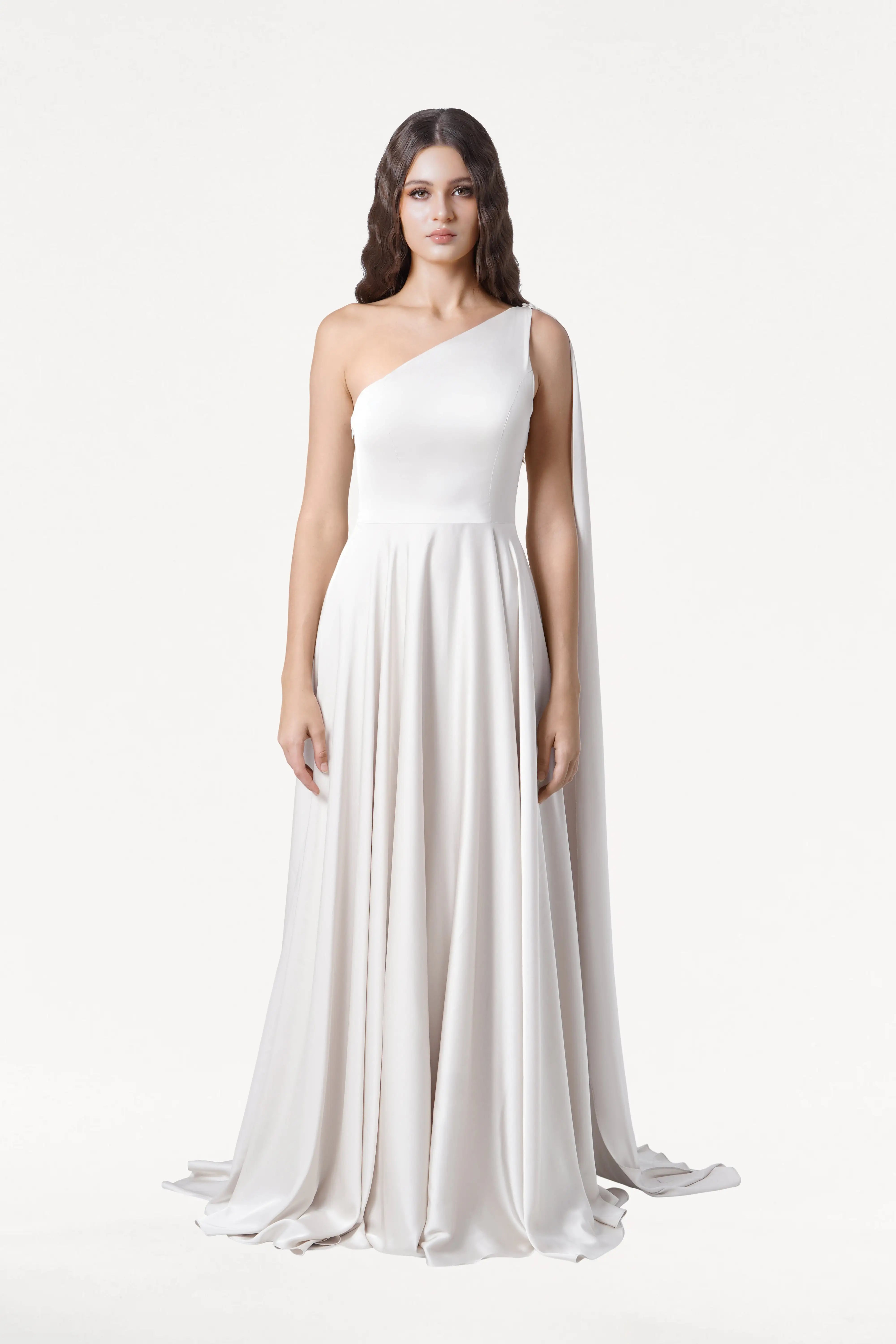 A woman with long, wavy dark hair and a neutral expression models the Lyra Dress in Ivory Satin by TH&TH—a size-inclusive, flowing, one-shoulder gown with a cape-like drape—against a plain white background.