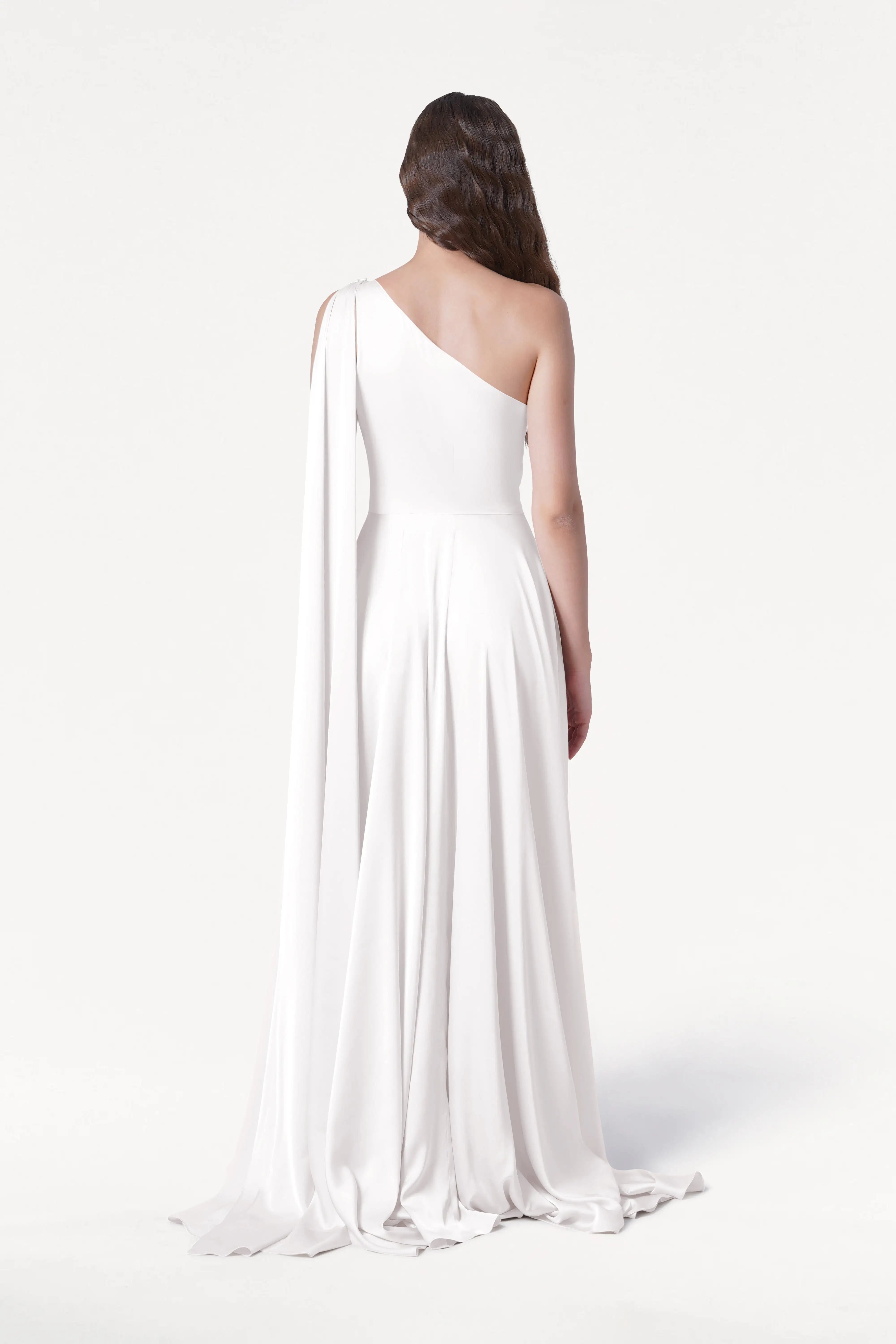 A woman with long wavy hair stands facing away, wearing the TH&TH Lyra Dress in Ivory Satin, a size-inclusive, flowing one-shoulder gown with a draped floor-length skirt against a plain white background.