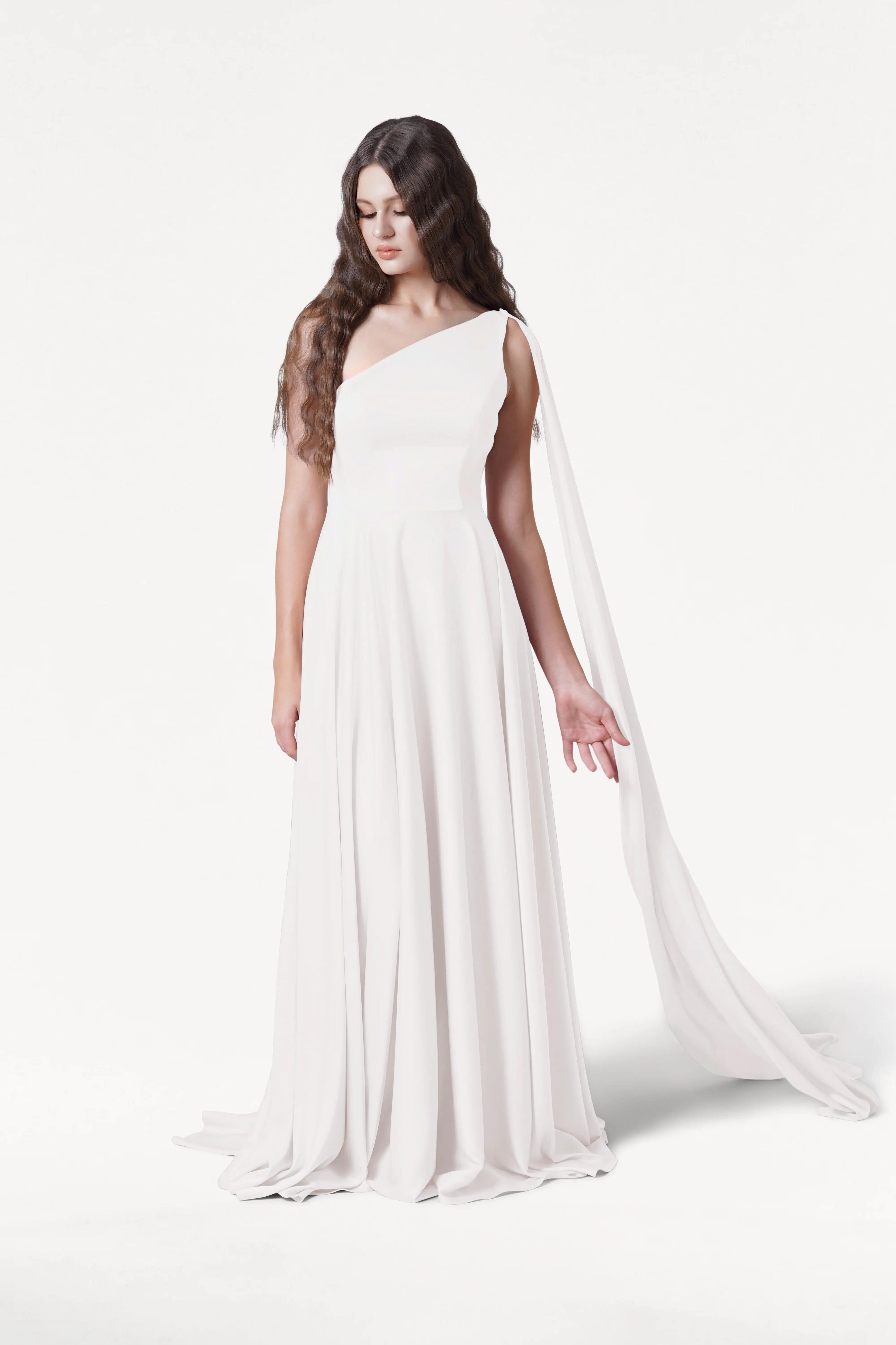A woman with long, wavy brown hair models the size-inclusive Lyra Dress in Ivory Satin by TH&TH. The flowing one-shoulder gown features a draped sash. She stands against a plain white background, looking down with relaxed pose.