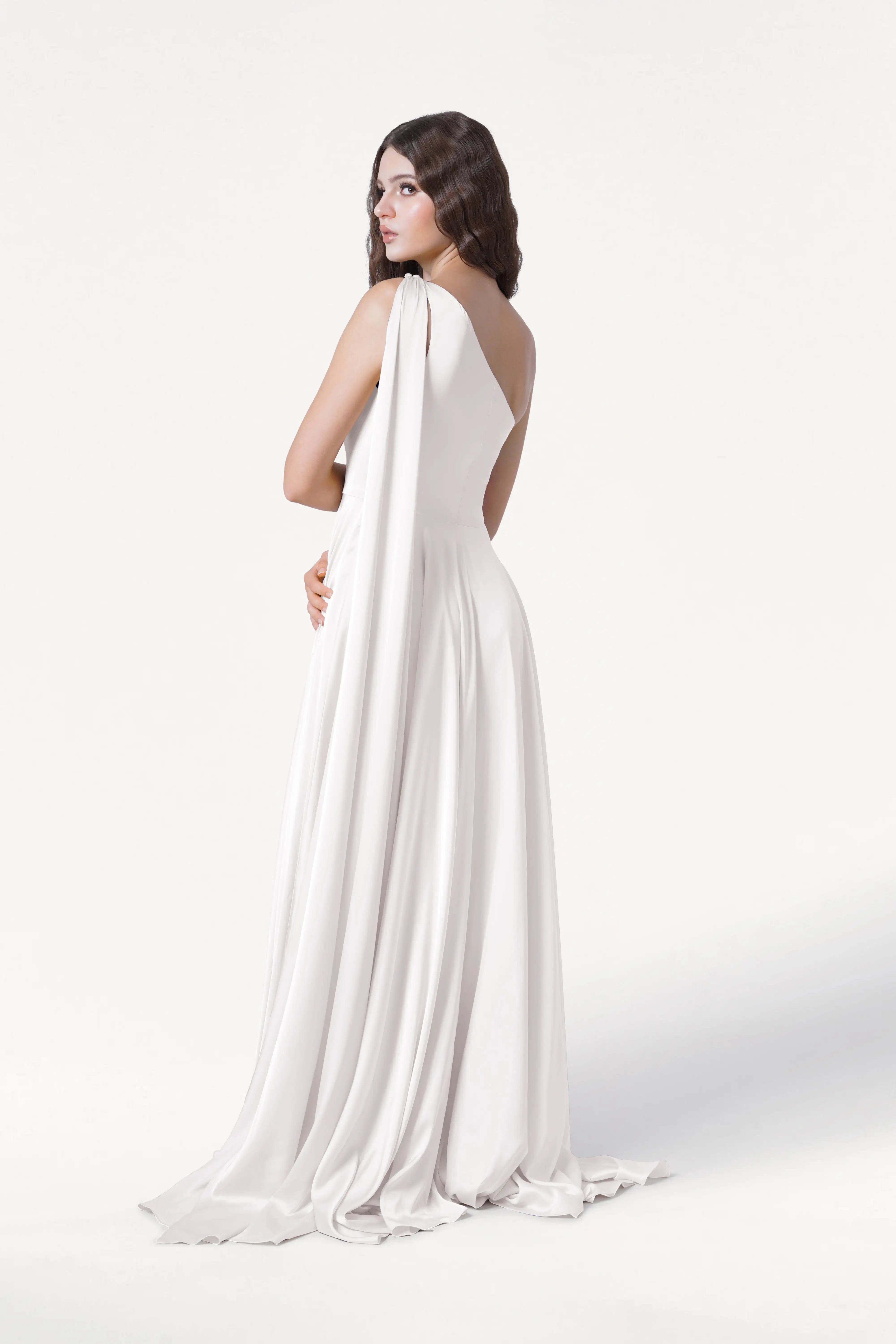 A woman with long dark hair in loose waves models the TH&TH Lyra Dress in Ivory Satin—a sleeveless, floor-length bridesmaid dress—as she stands against a plain white background, turned slightly away and looking over her shoulder.