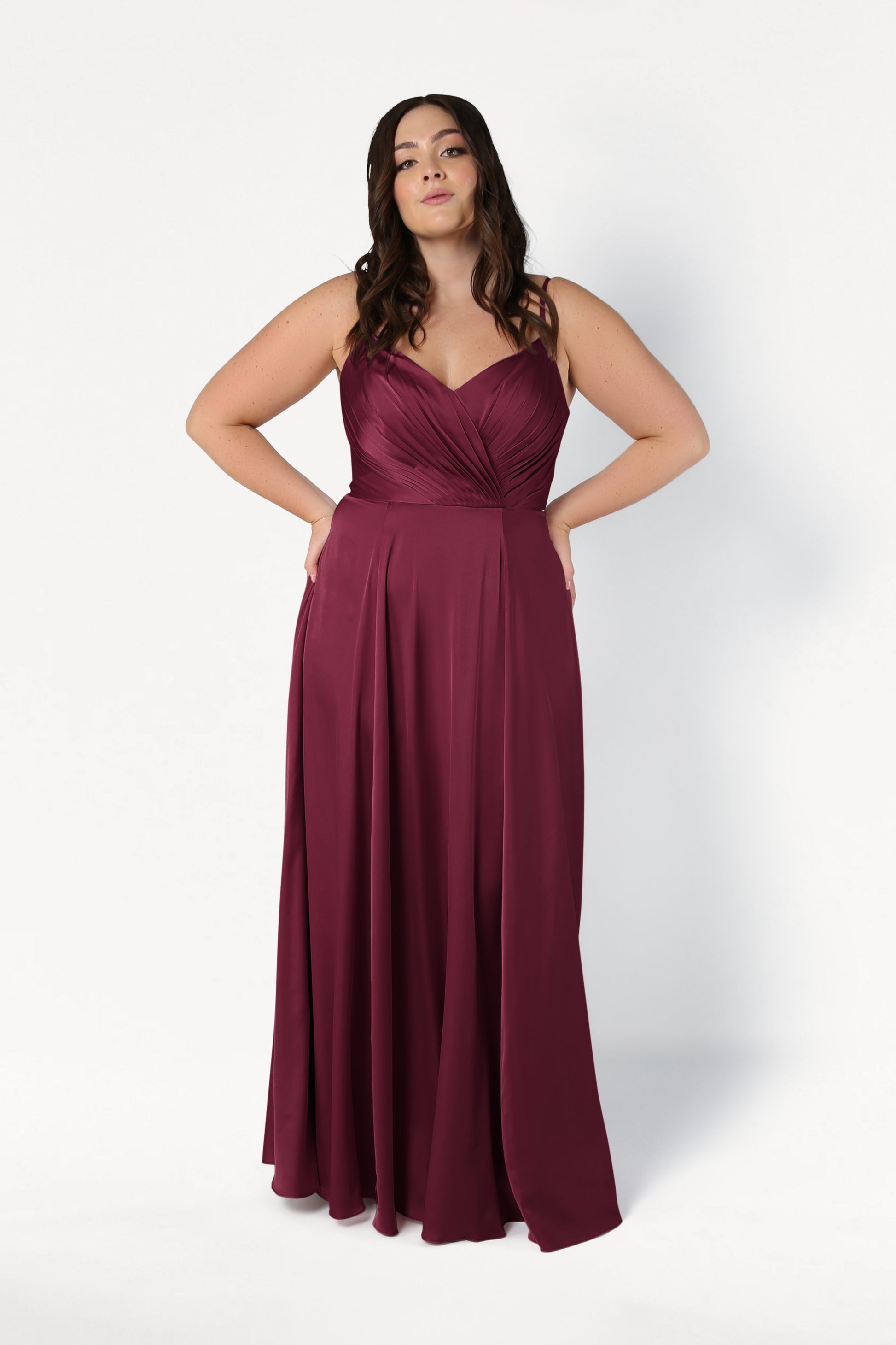 A plus-size model wears the TH&TH Isla Bridesmaid Dress in Merlot Satin, featuring a pleated sweetheart bodice, spaghetti straps, and a flowing floor-length A-line skirt, posing against a white background.